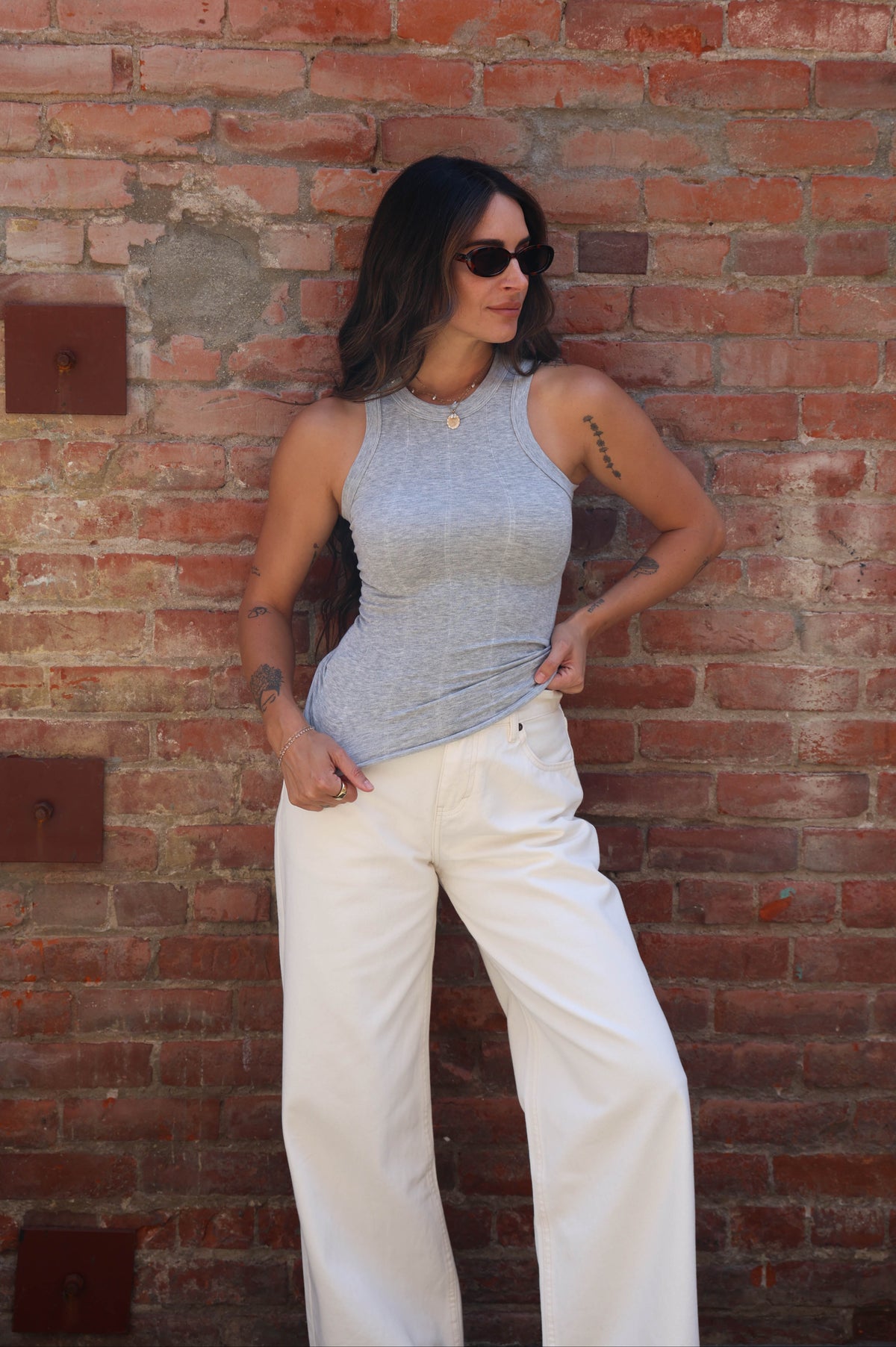 A woman wearing a gray sleeveless top and white wide-leg pants stands against a red brick wall.