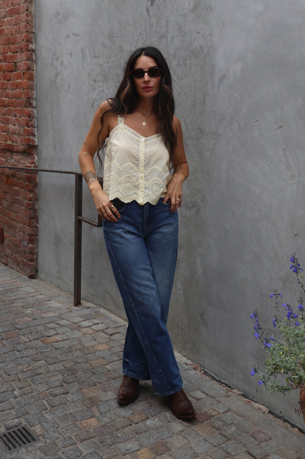 Woman in a white lace top and blue jeans standing against a gray wall.
