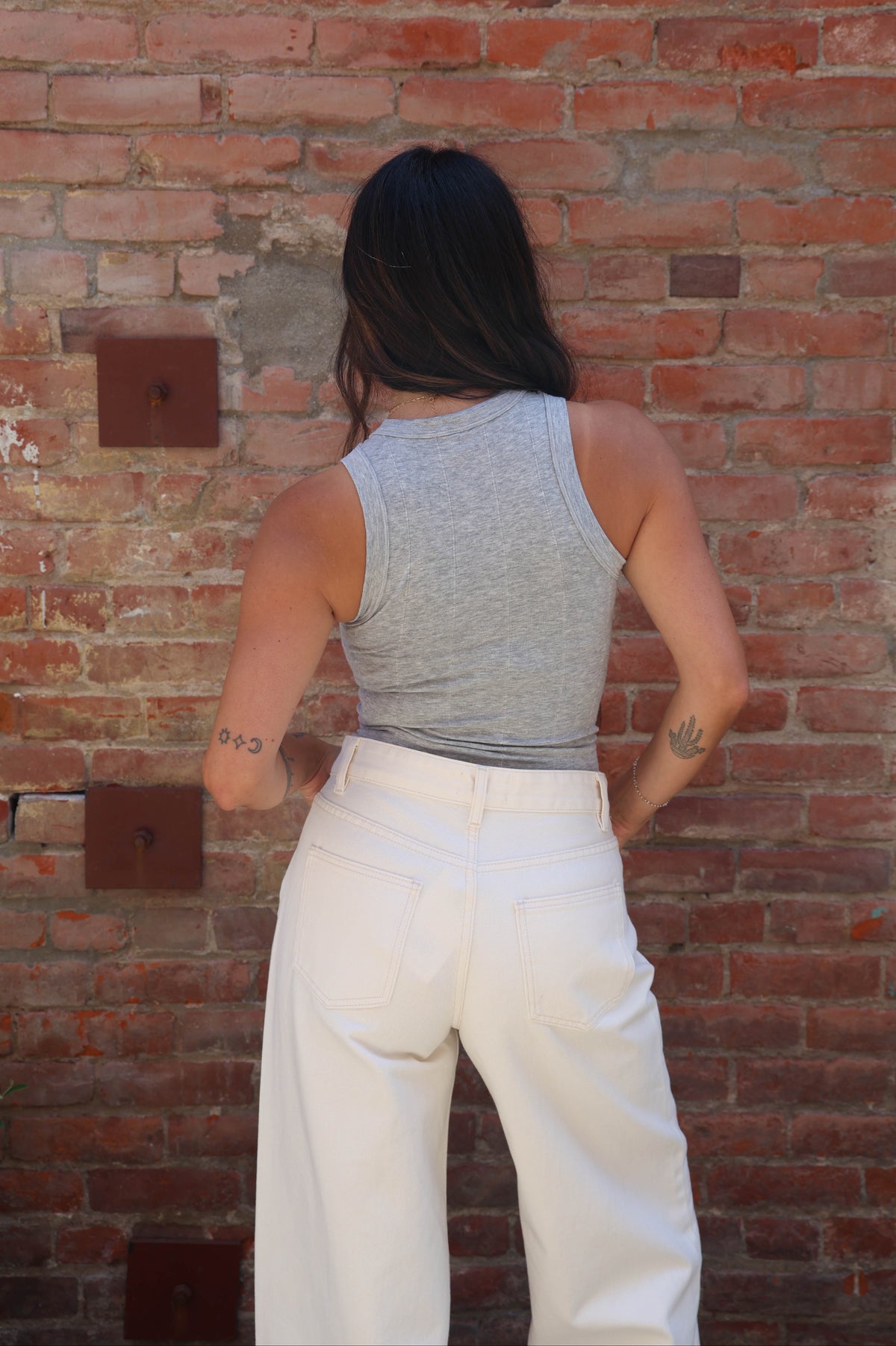 A woman viewed from behind wearing a heather gray ribbed tank top and white wide-leg pants, standing against a red brick wall.