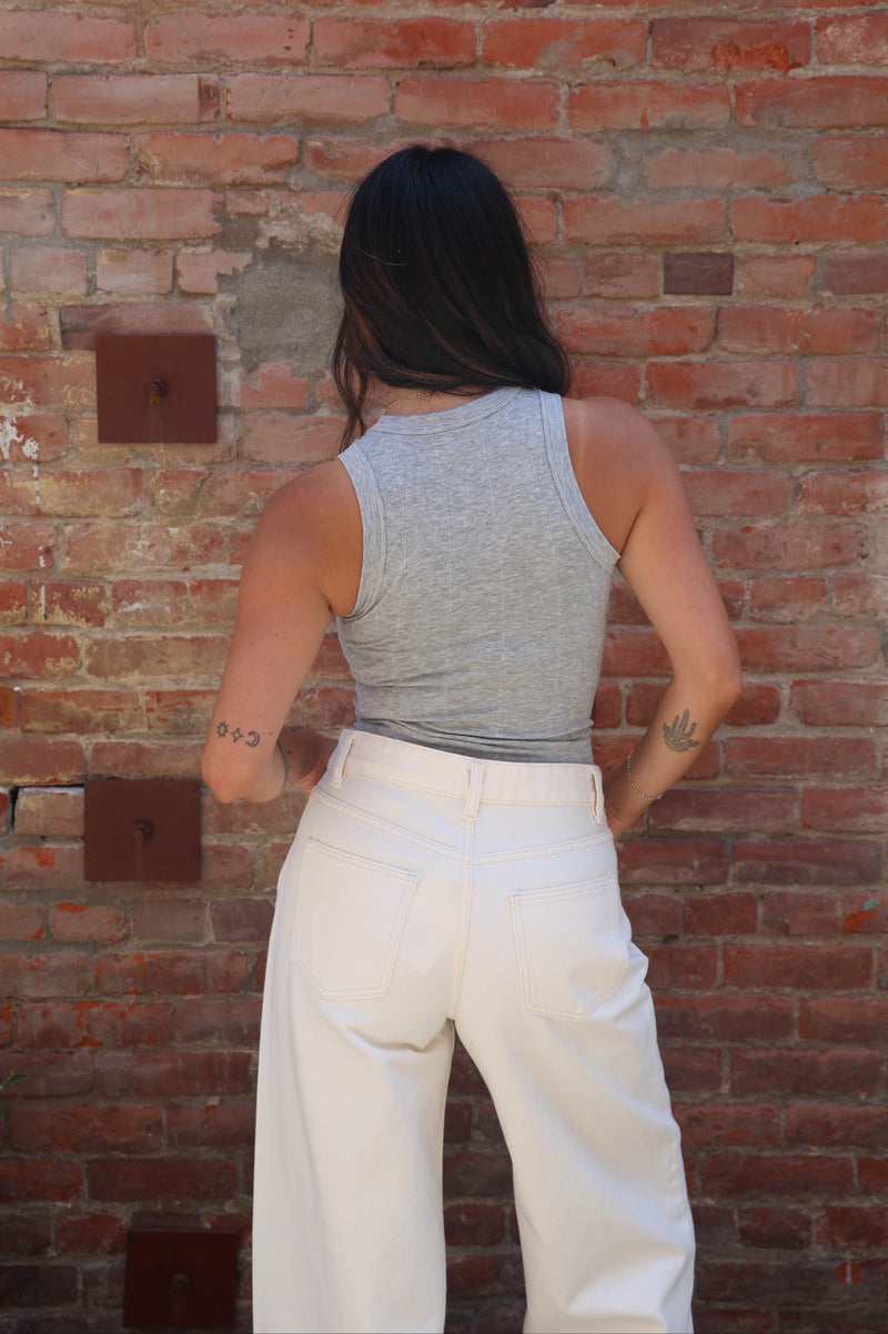 A woman viewed from behind wearing a heather gray ribbed tank top and white wide-leg pants, standing against a red brick wall.