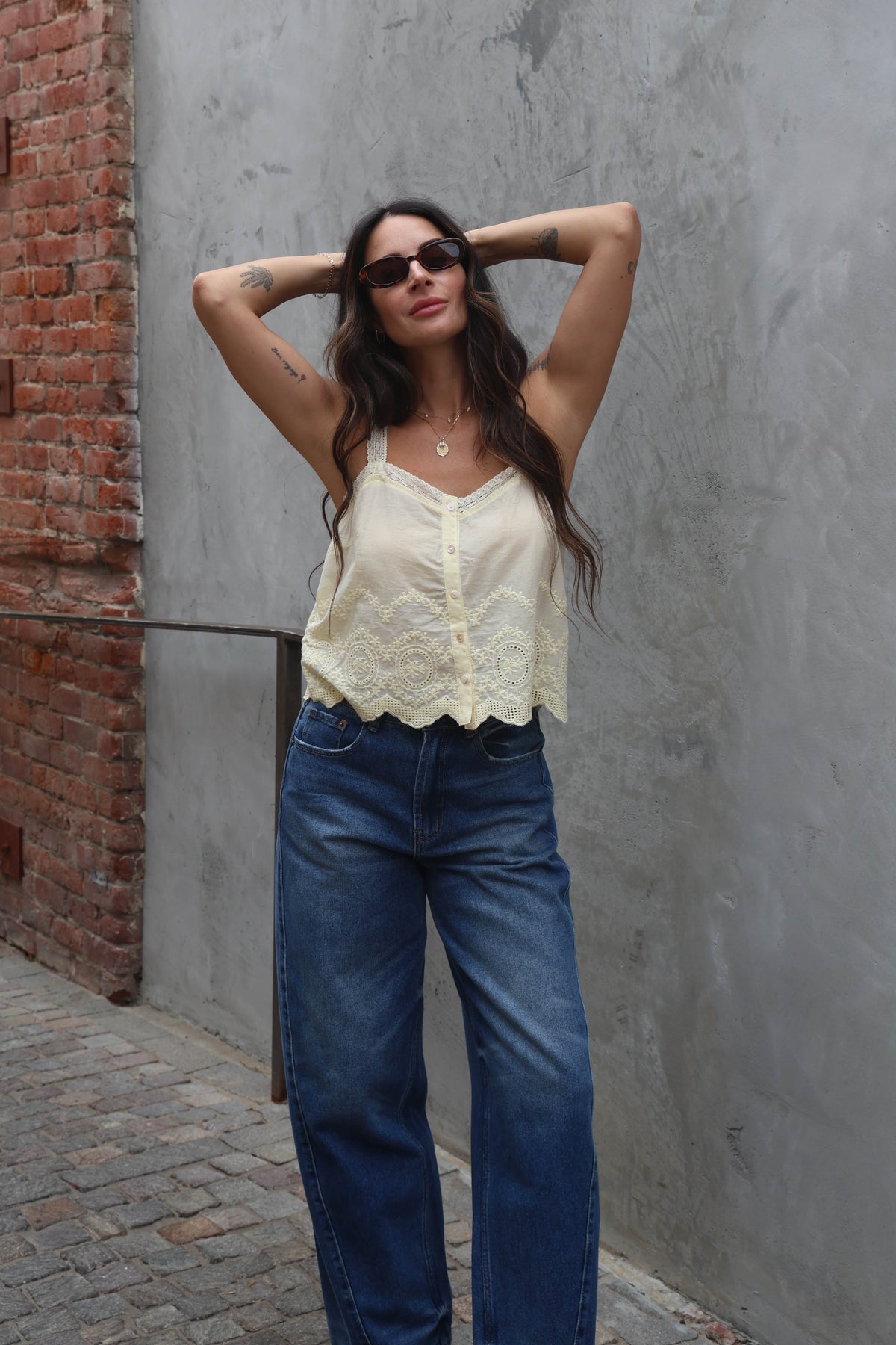 Woman wearing a white lace top and blue jeans against a gray wall.