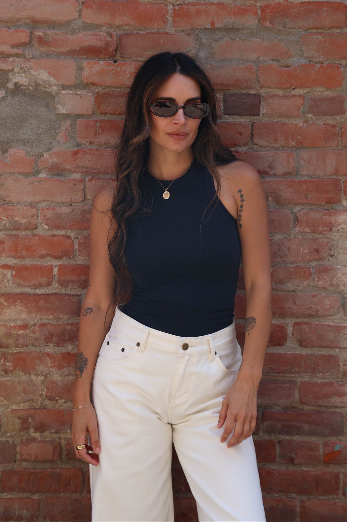 A woman with long dark hair stands against a red brick wall wearing a sleeveless black ribbed top, white high-waisted cropped pants, and dark sunglasses.
