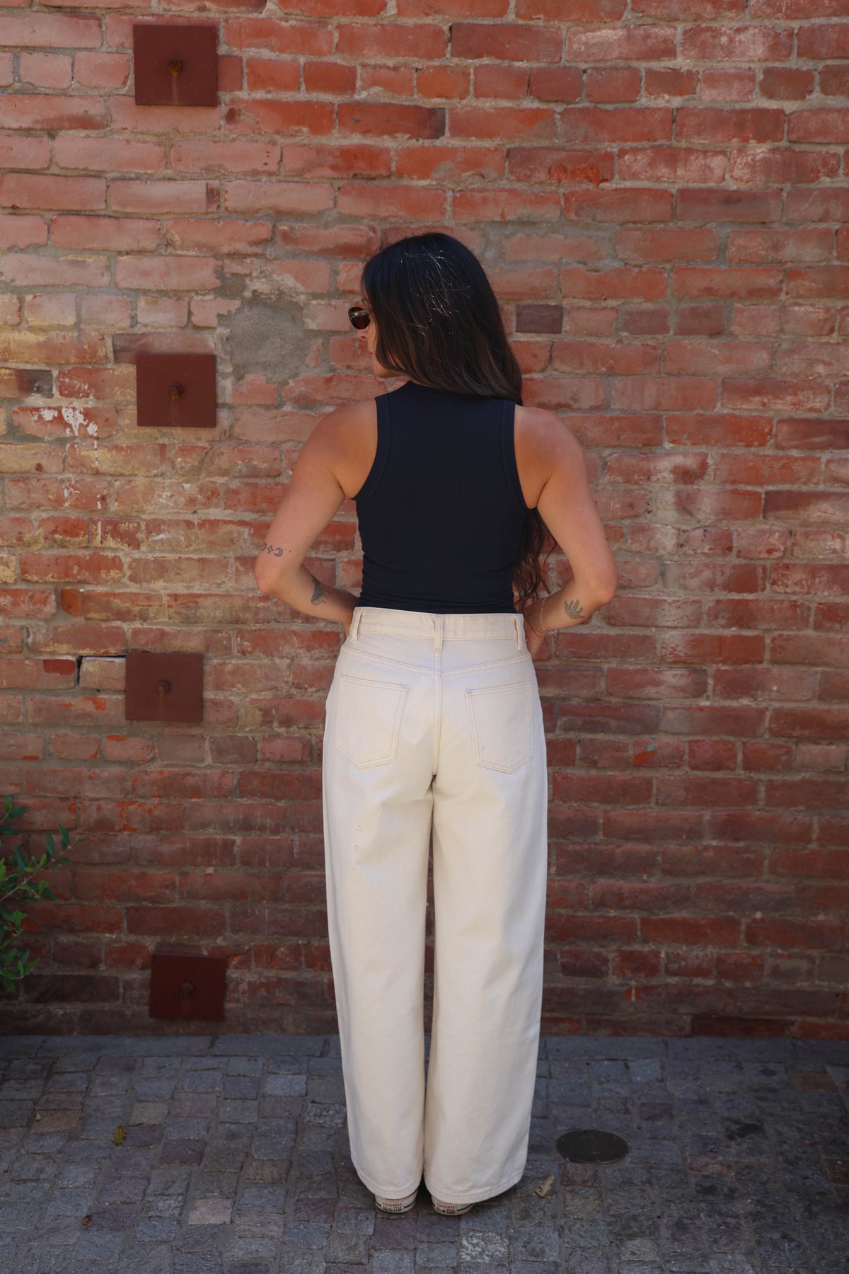 A woman stands with her back to the camera wearing a black sleeveless top and high-waisted, wide-leg cream trousers, posing in front of a red brick wall.