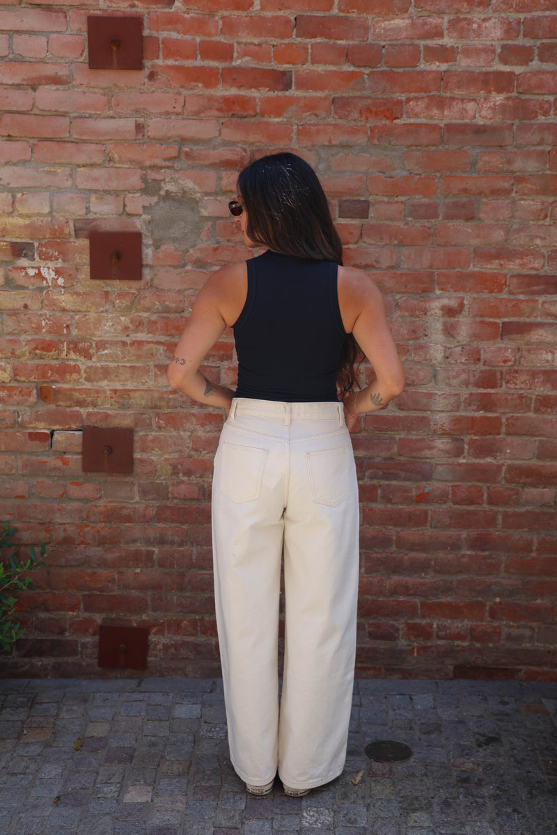 A woman stands with her back to the camera wearing a black sleeveless top and high-waisted, wide-leg cream trousers, posing in front of a red brick wall.