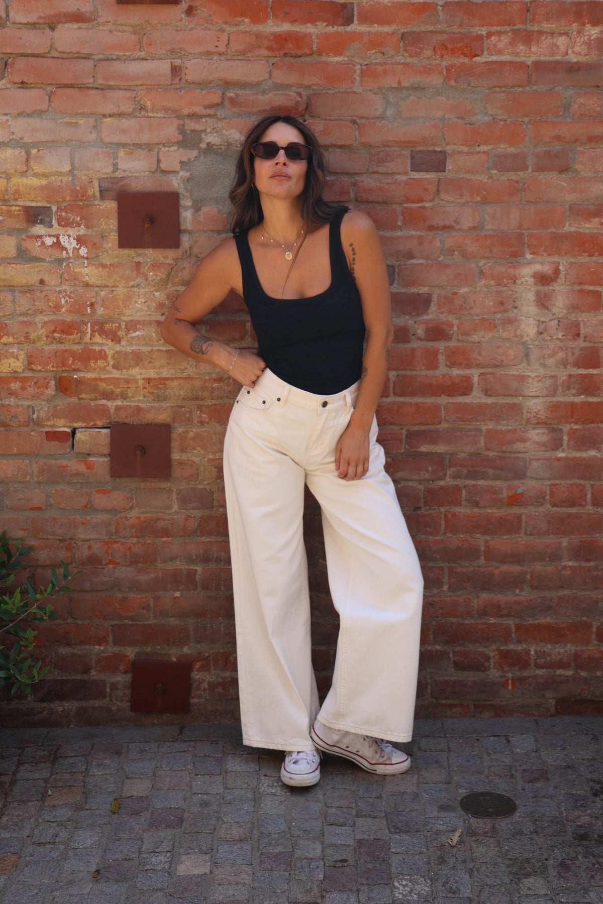 A woman posing in front of a red brick wall, wearing a black tank top, off-white wide-leg pants, and white sneakers.