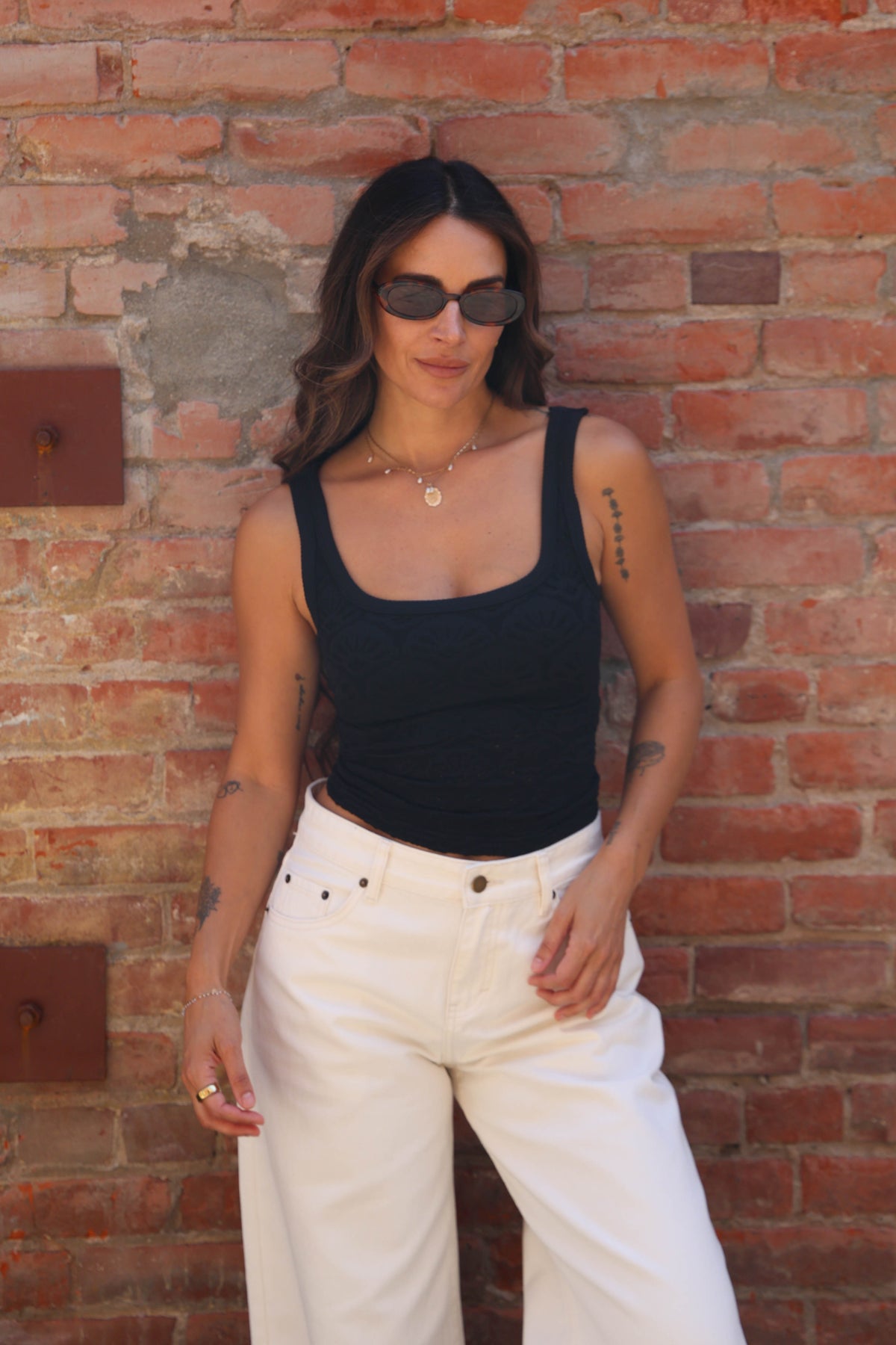 A woman wearing a black ribbed tank top and white wide-leg jeans stands against a red brick wall.