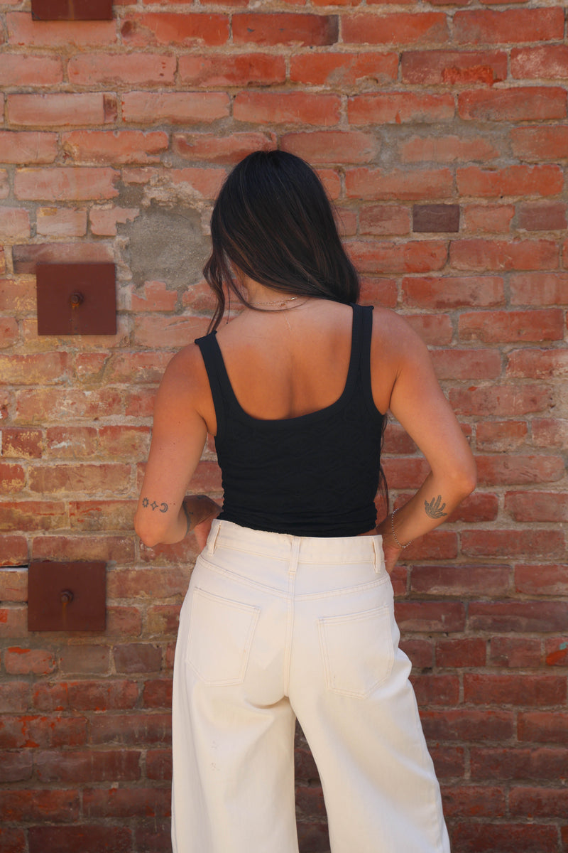 A woman viewed from behind wearing a black tank top and white wide-leg pants, standing against a red brick wall.