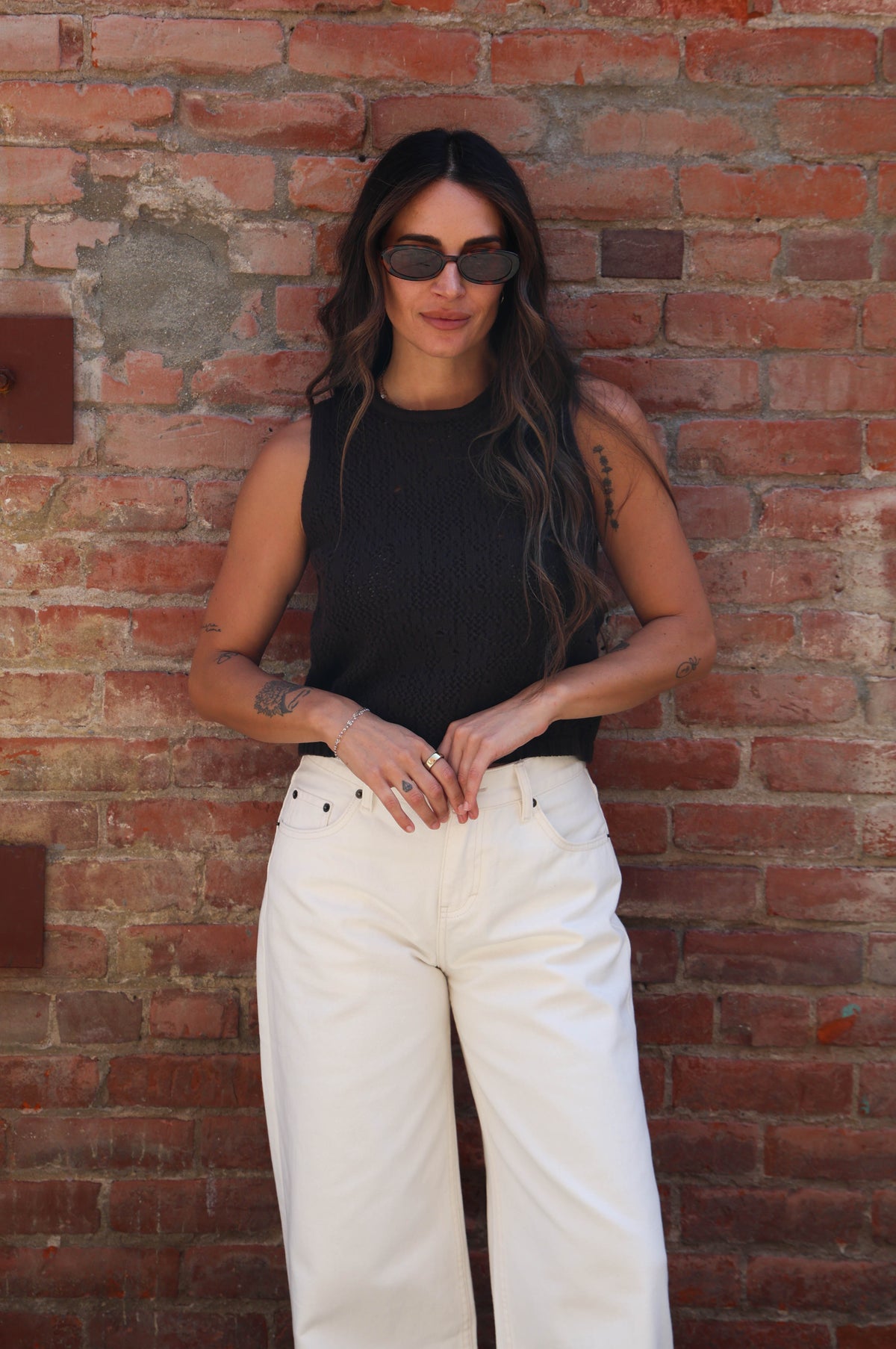 A woman wearing a black sleeveless top and off-white wide-leg pants stands against a red brick wall.