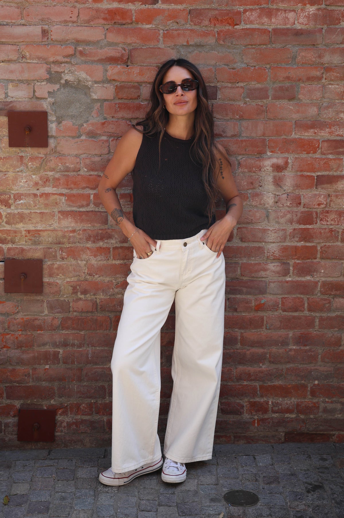 A woman stands against a red brick wall wearing a black sleeveless top, off-white wide-leg pants, and white sneakers.