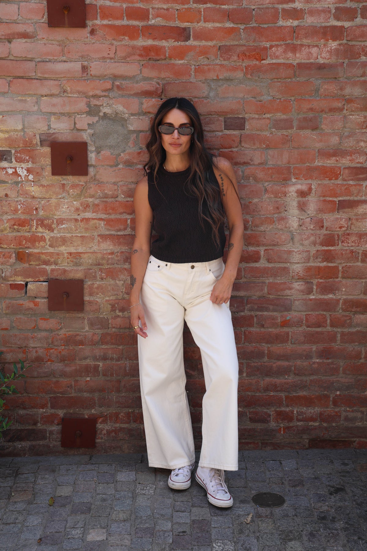 A woman stands against a red brick wall wearing a black sleeveless top, off-white wide-leg pants, and white sneakers.