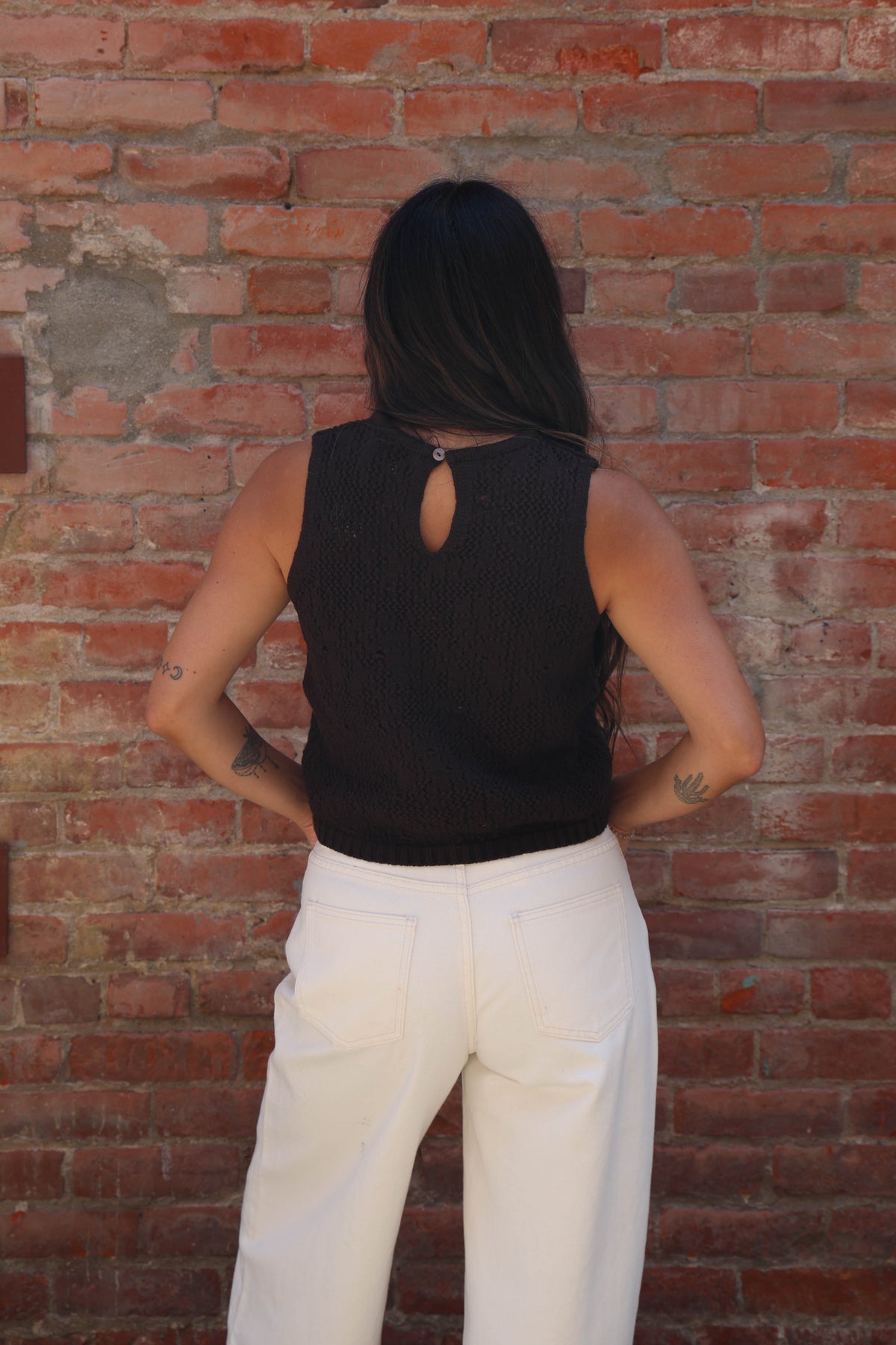 A woman viewed from behind wearing a black sleeveless ribbed top with a keyhole cutout and white pants, standing against a red brick wall.
