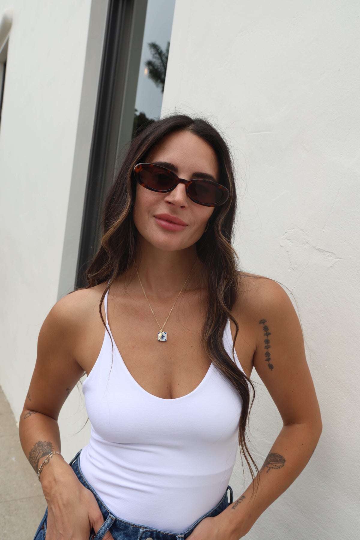 Woman wearing sunglasses and a white tank top against a light-colored wall.