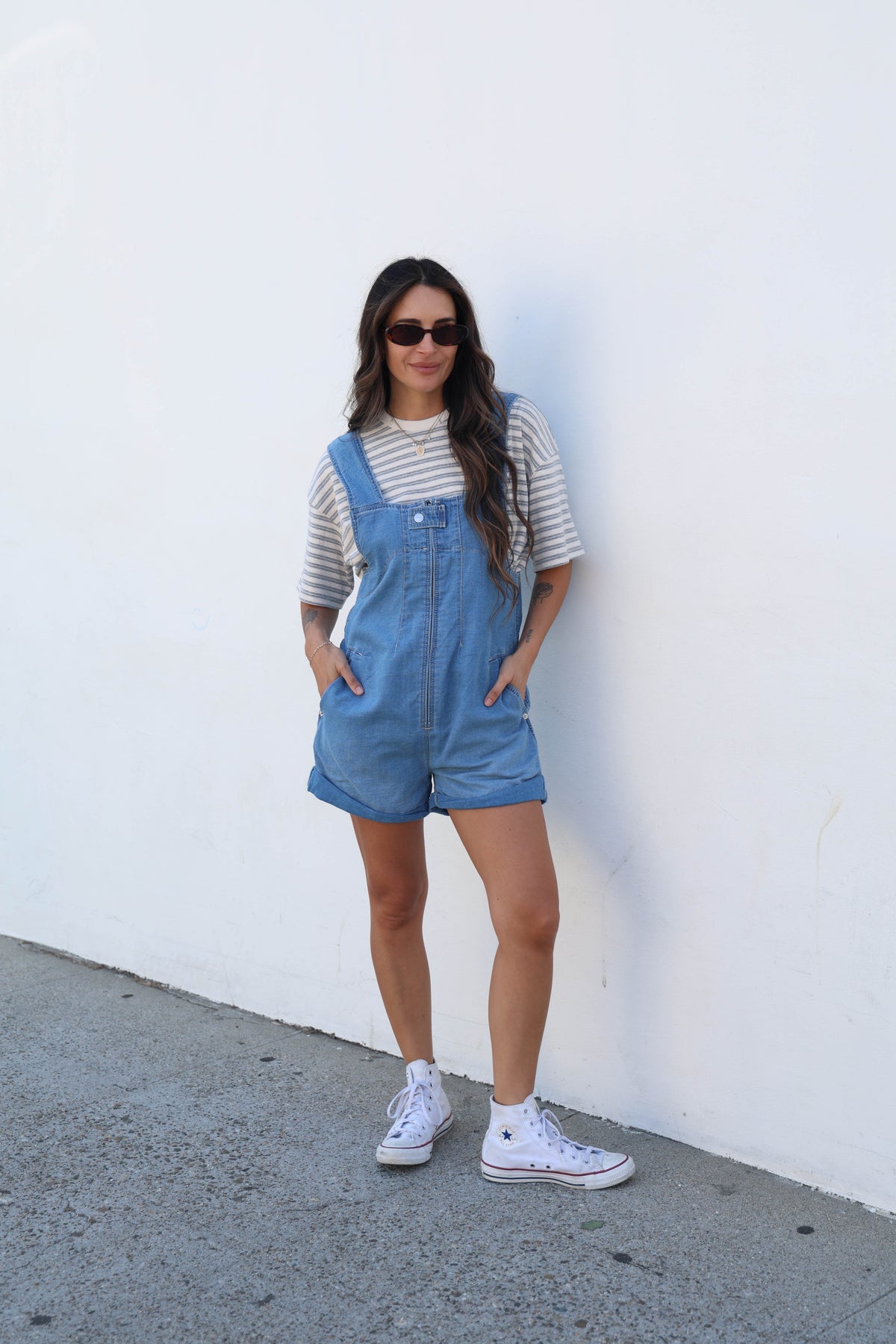 A woman wearing a light blue denim overall romper over a gray and white striped t-shirt, styled with white sneakers, standing against a plain white wall.