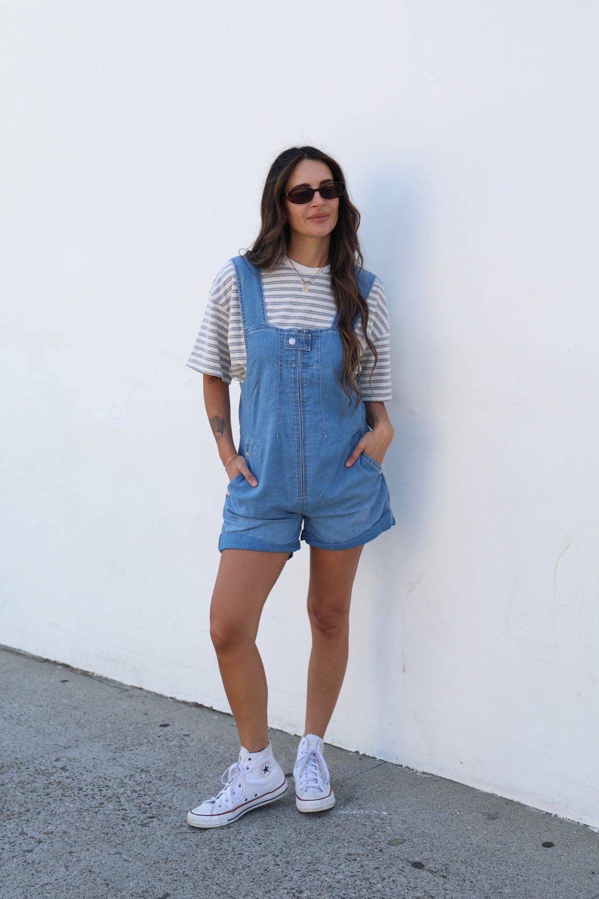 A woman stands against a white wall wearing blue denim overall shorts over a white and gray striped t-shirt, styled with white sneakers and sunglasses.
