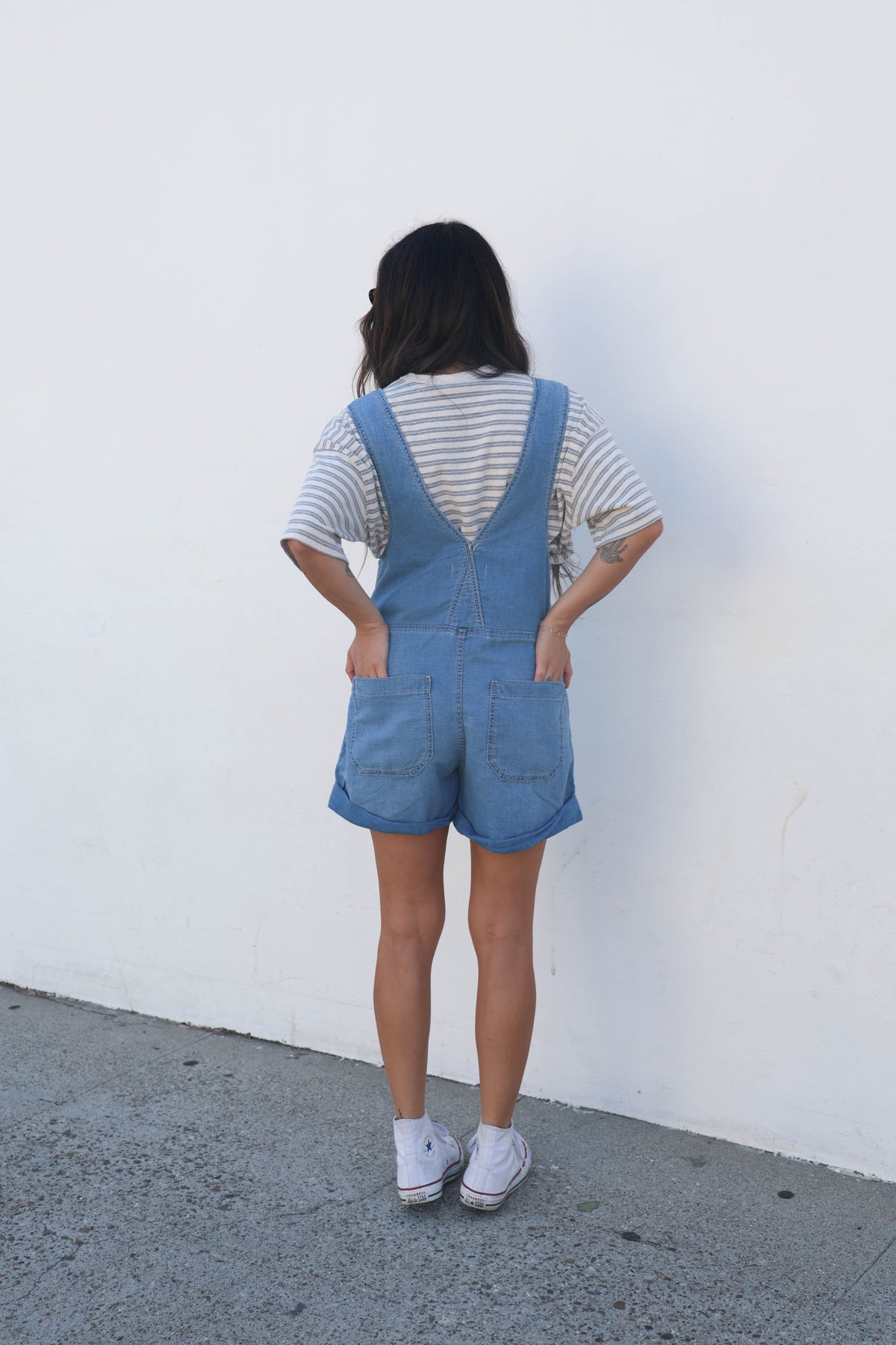 A person standing with their back to the camera, wearing light blue denim overall shorts over a white and gray striped t-shirt, paired with white sneakers, set against a plain white wall.
