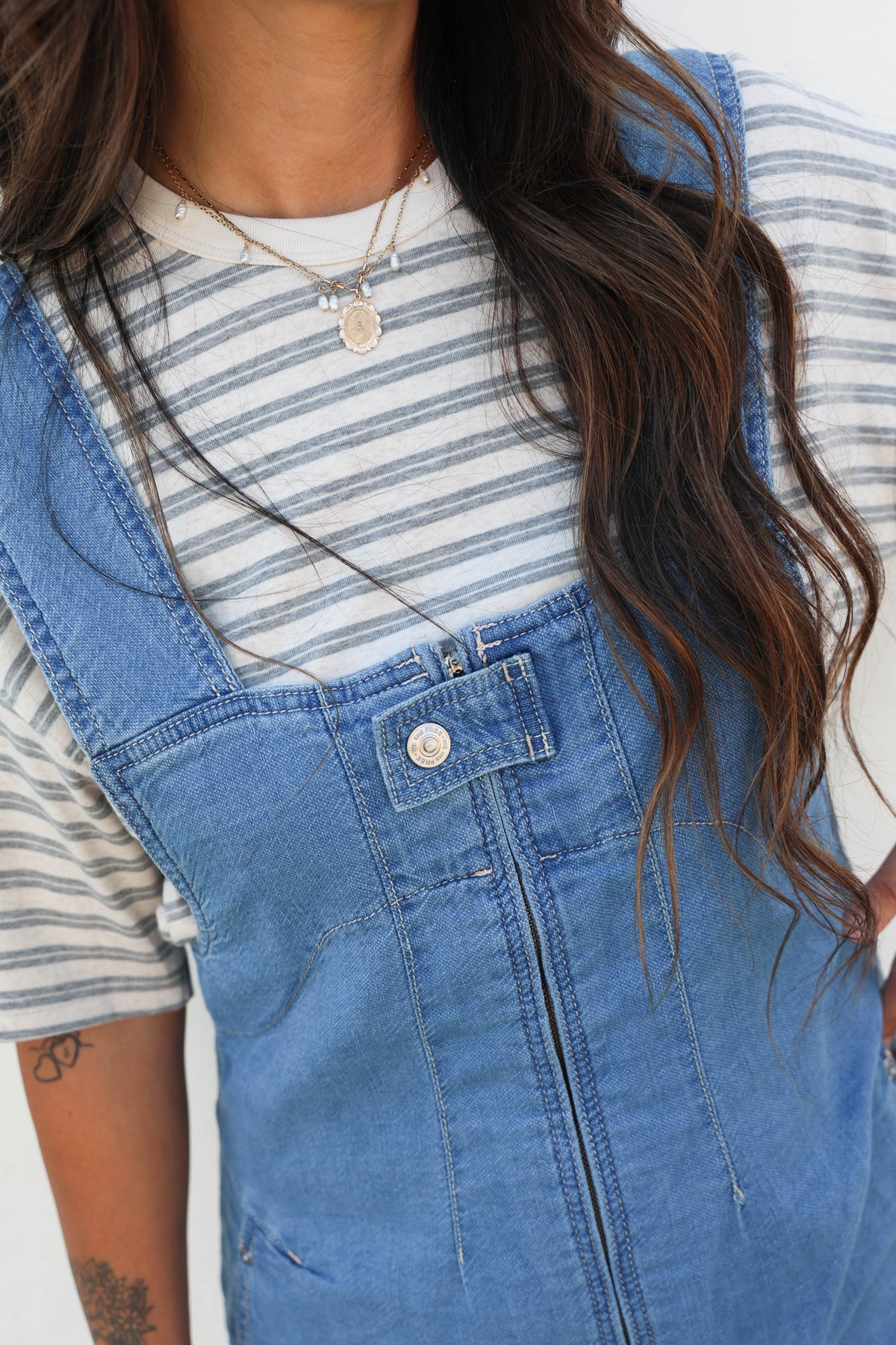 A close-up of a person wearing blue denim overall-style shorts over a white and gray striped t-shirt, with long wavy brown hair and a silver necklace visible.