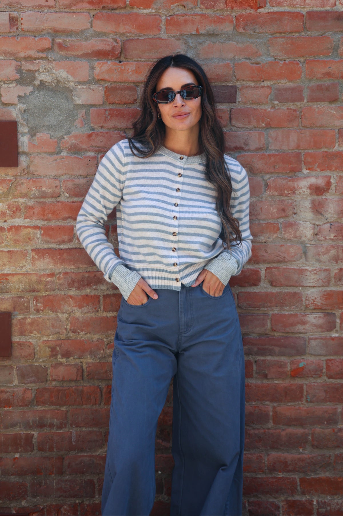 A woman wearing a light blue and white striped long-sleeve button-up top and blue wide-leg pants, standing against a red brick wall.