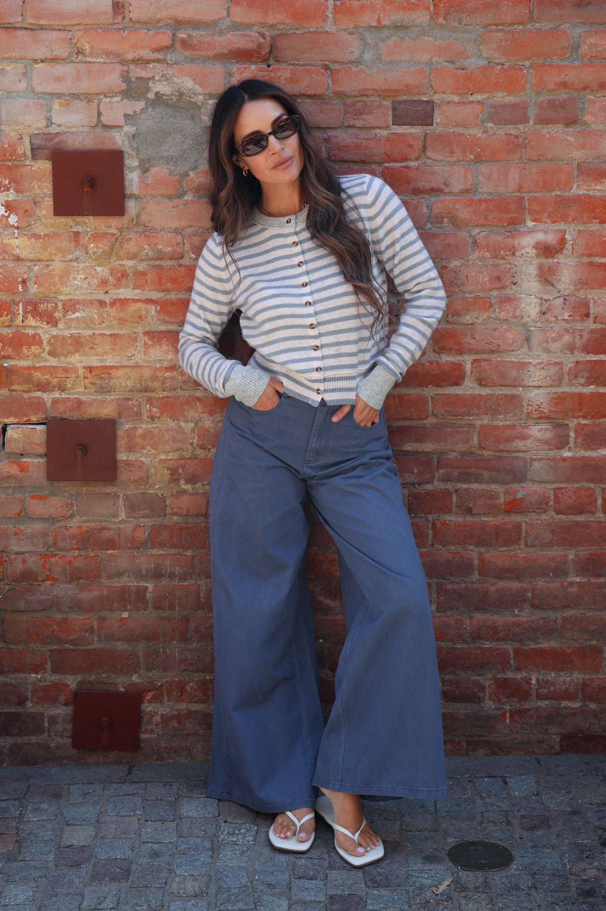 A woman wearing a striped long-sleeve button-up shirt and wide-leg blue pants stands against a red brick wall.