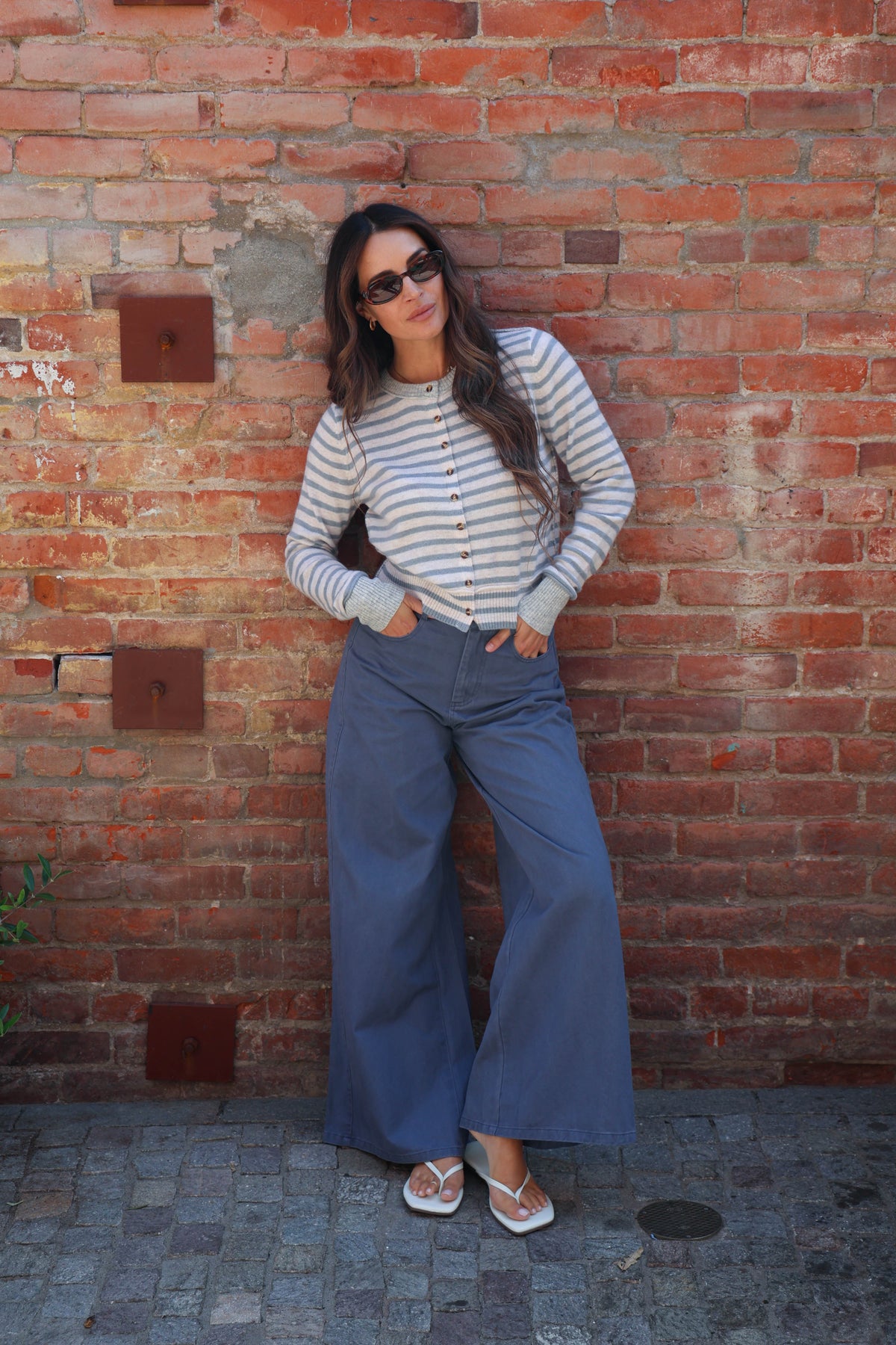 A woman wearing a striped long-sleeve button-up shirt and wide-leg blue pants stands against a red brick wall.