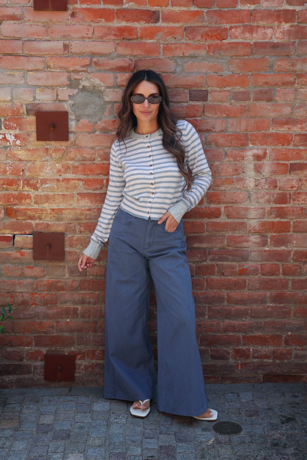 A woman stands against a red brick wall wearing a light gray and white striped long-sleeve button-up shirt, wide-leg blue pants, and white slide sandals.
