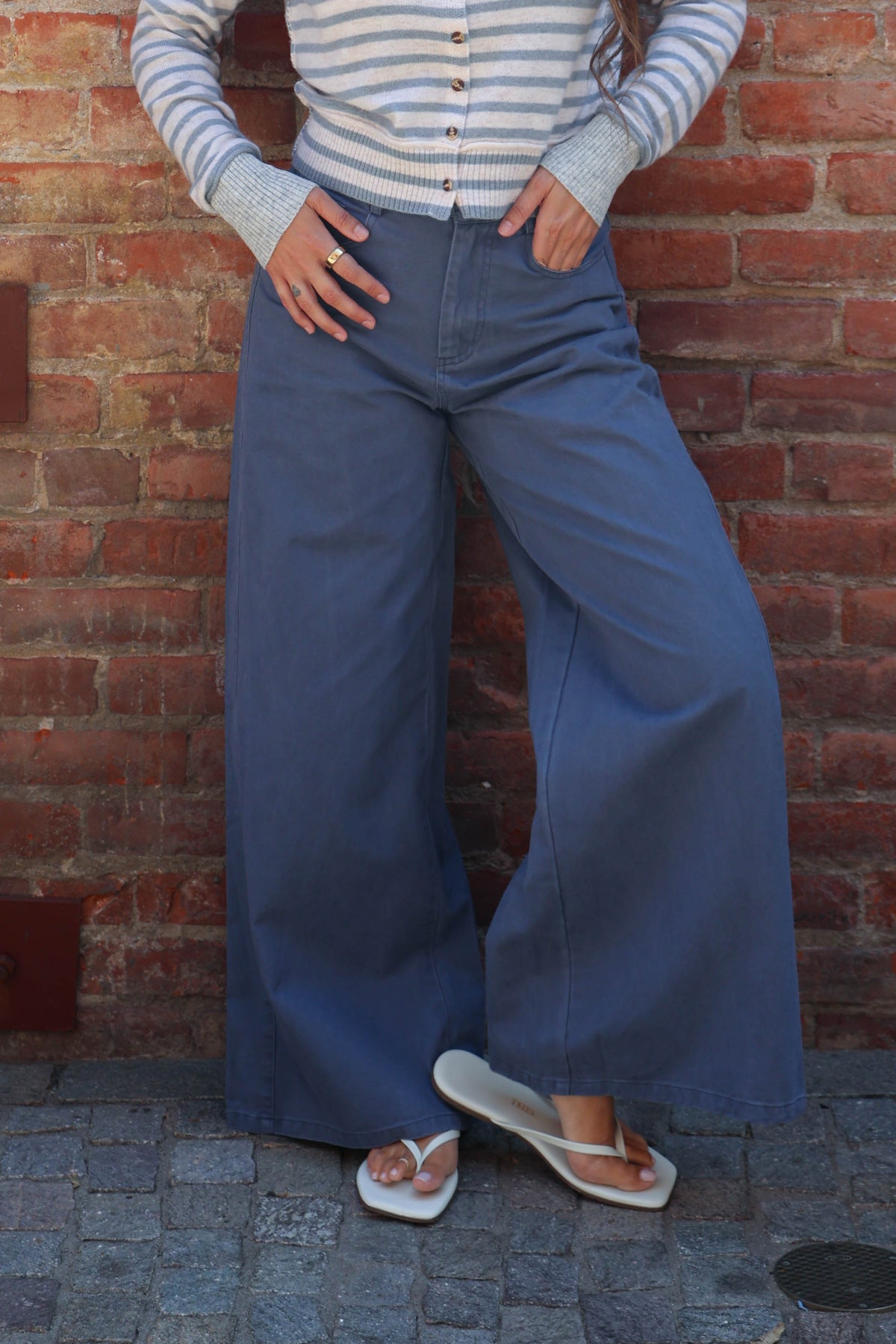 A person wearing wide-leg blue denim pants, a light blue and white striped long-sleeve top, and white flip-flop sandals, standing against a red brick wall.