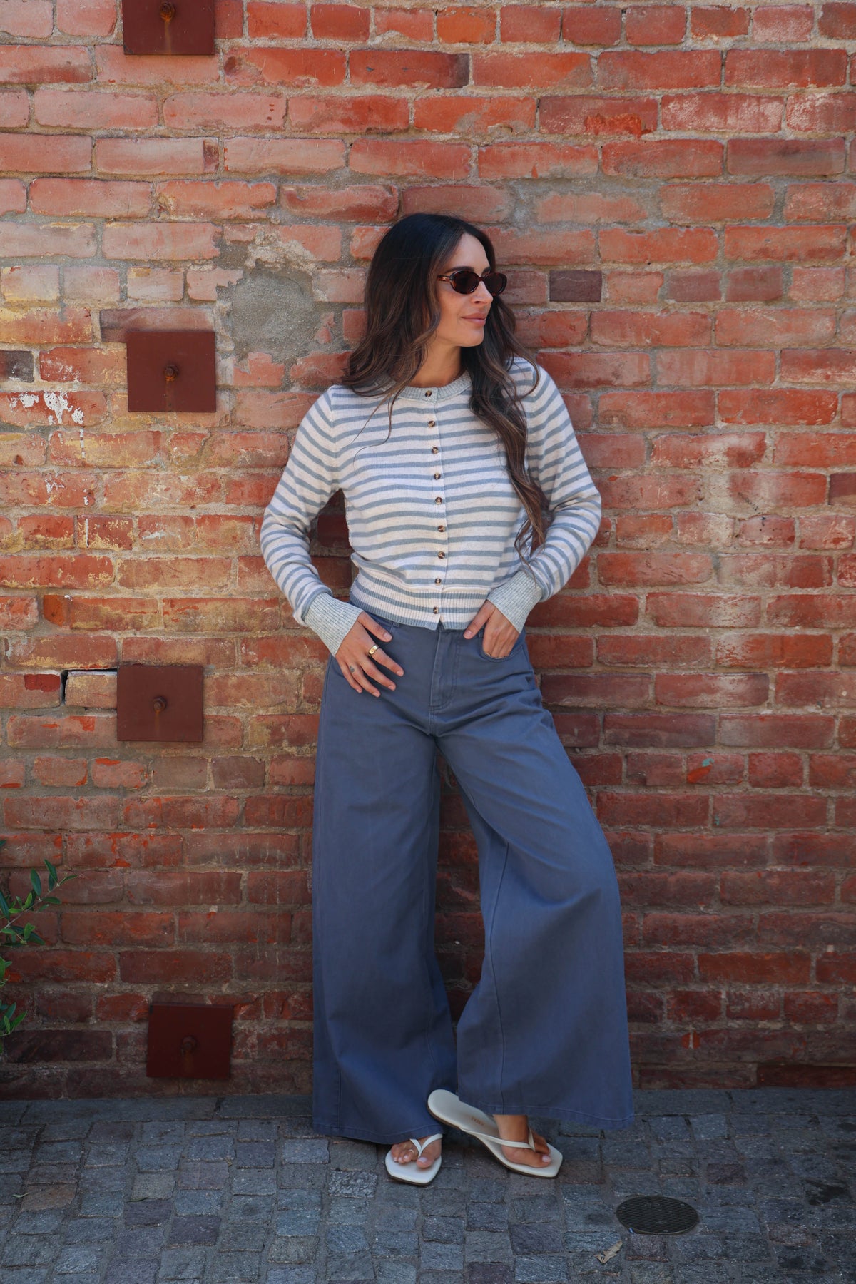 A woman stands against a red brick wall wearing a cream and navy striped long-sleeve button-up top, wide-leg blue pants, and white slide sandals.
