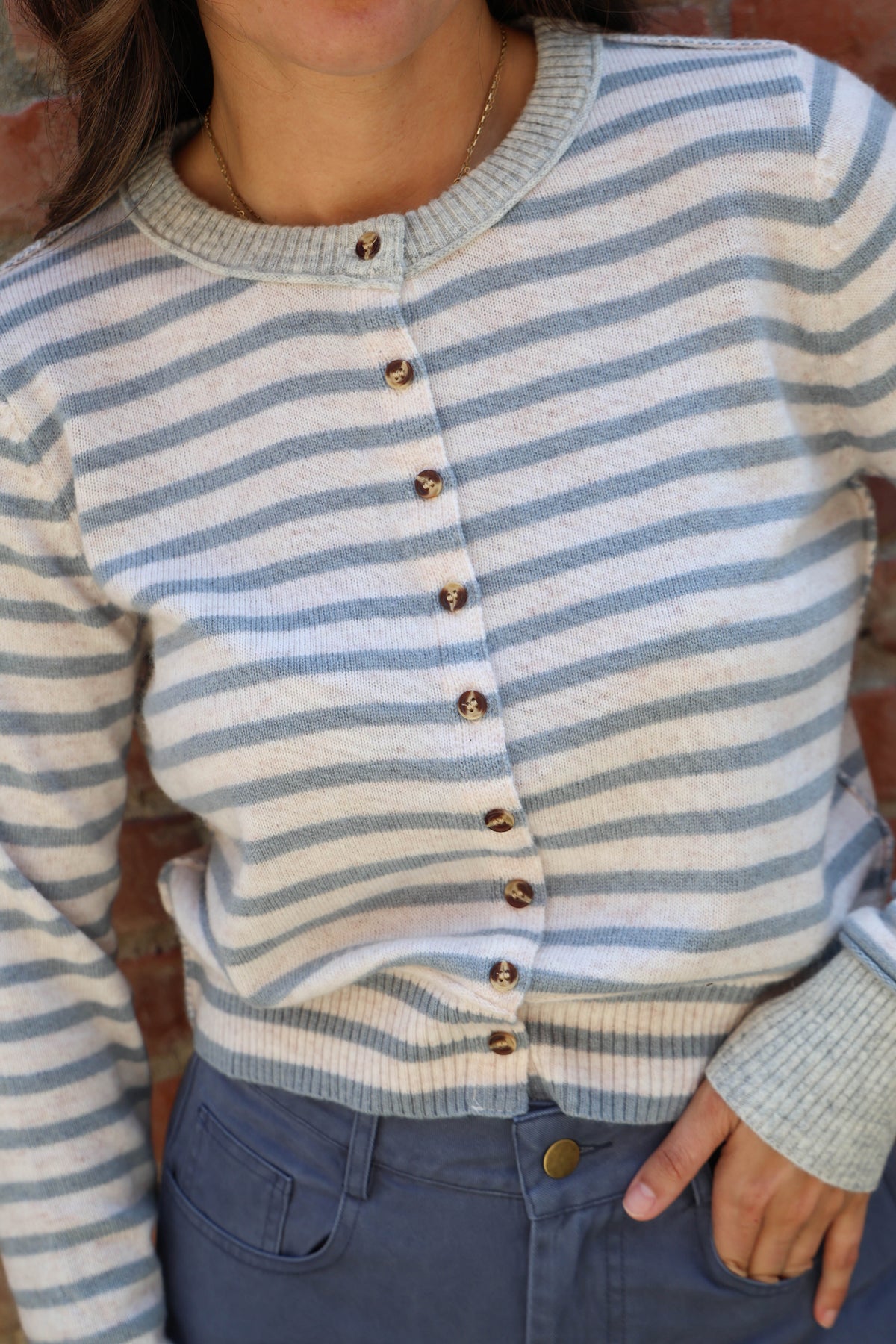 A close-up of a person wearing a light blue and off-white striped knit cardigan with brown buttons, paired with blue jeans.