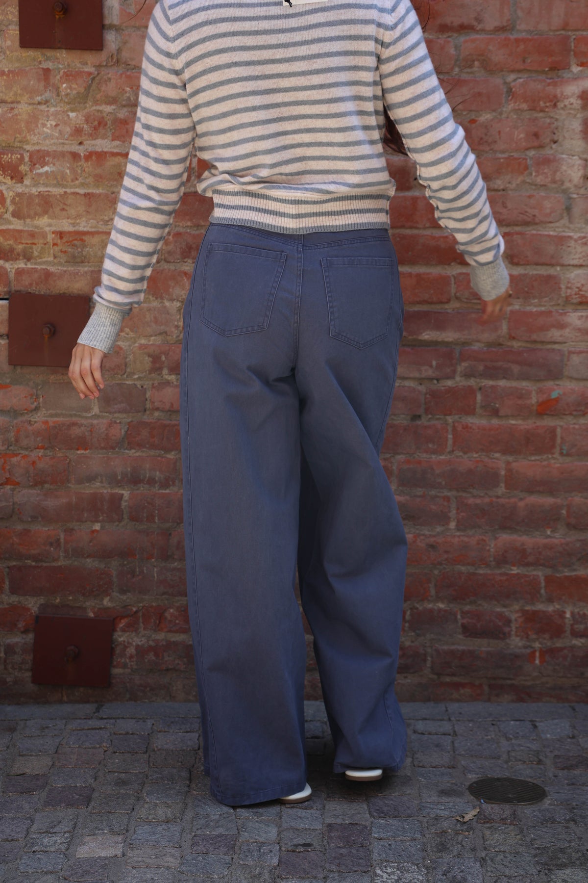 A person shown from behind wearing a cream and navy striped long-sleeve top and wide-leg blue jeans, standing against a red brick wall.