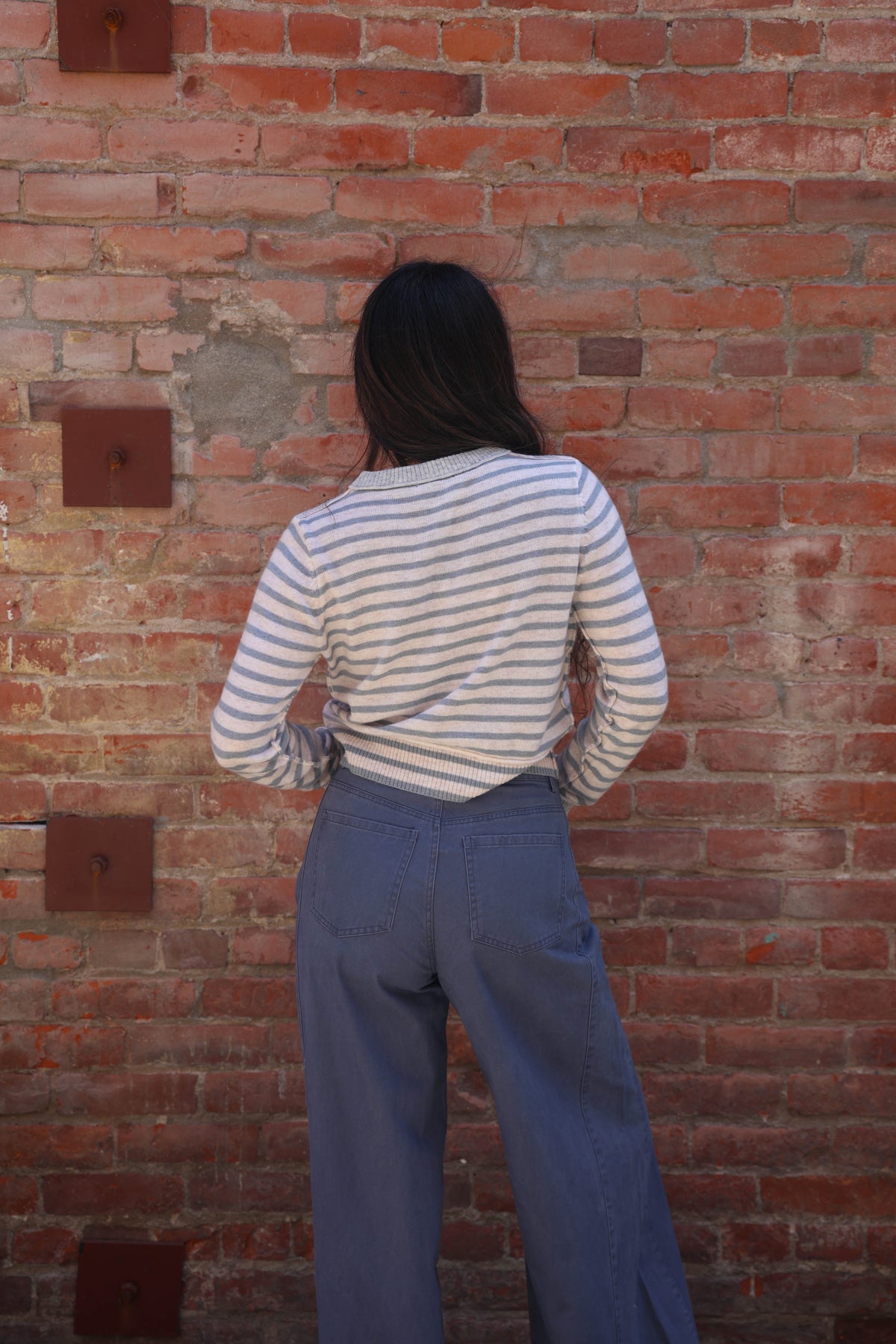 A person viewed from behind wearing a cream and navy blue horizontally striped long-sleeve shirt and blue wide-leg jeans, standing against a red brick wall.
