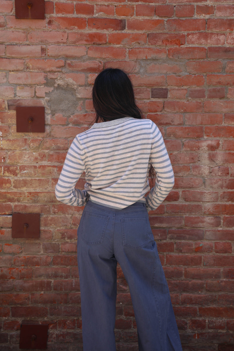 A person viewed from behind wearing a cream and navy blue horizontally striped long-sleeve shirt and blue wide-leg jeans, standing against a red brick wall.