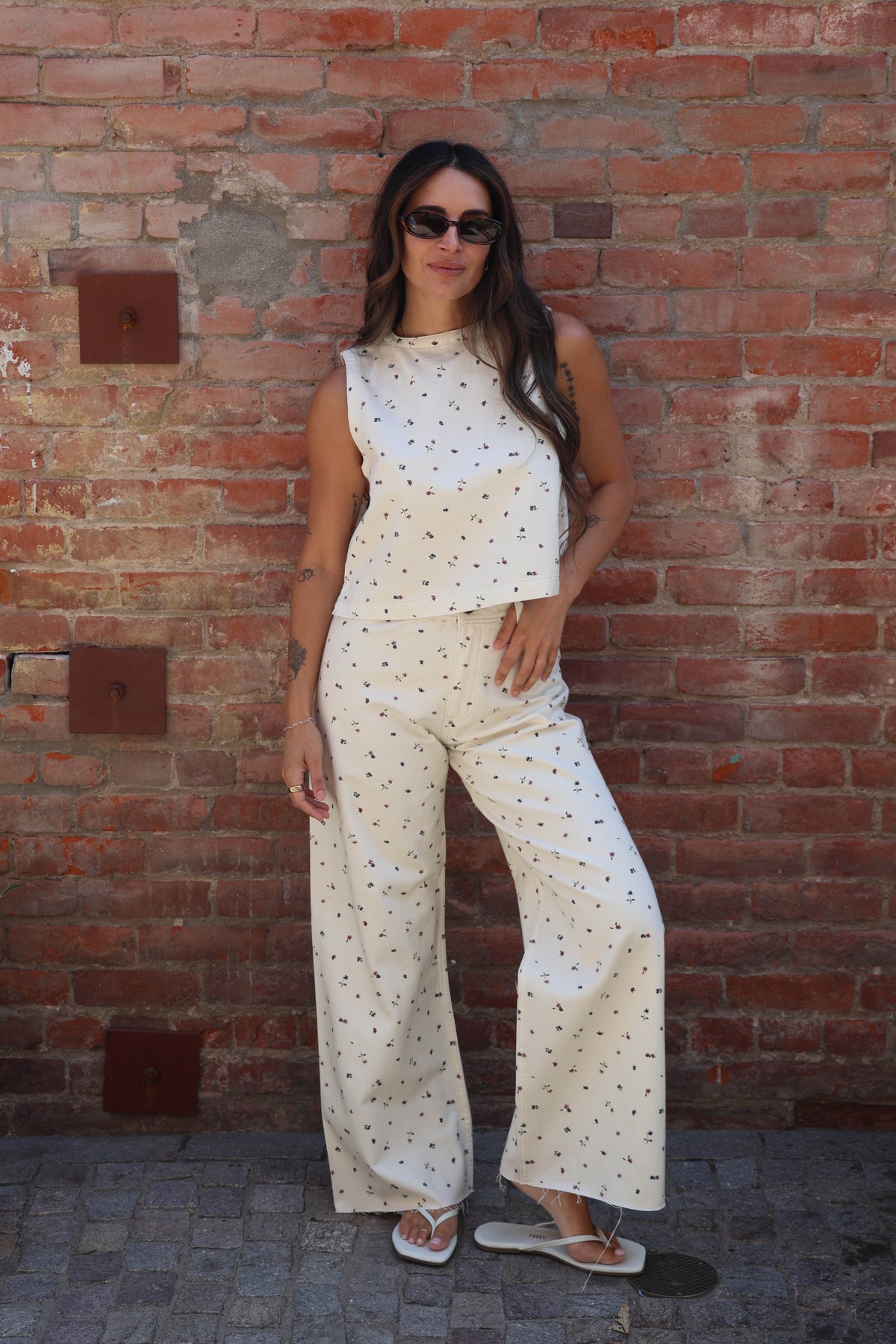 A woman wearing a matching sleeveless top and wide-leg pants set in an off-white fabric with small black polka dots, standing against a red brick wall.