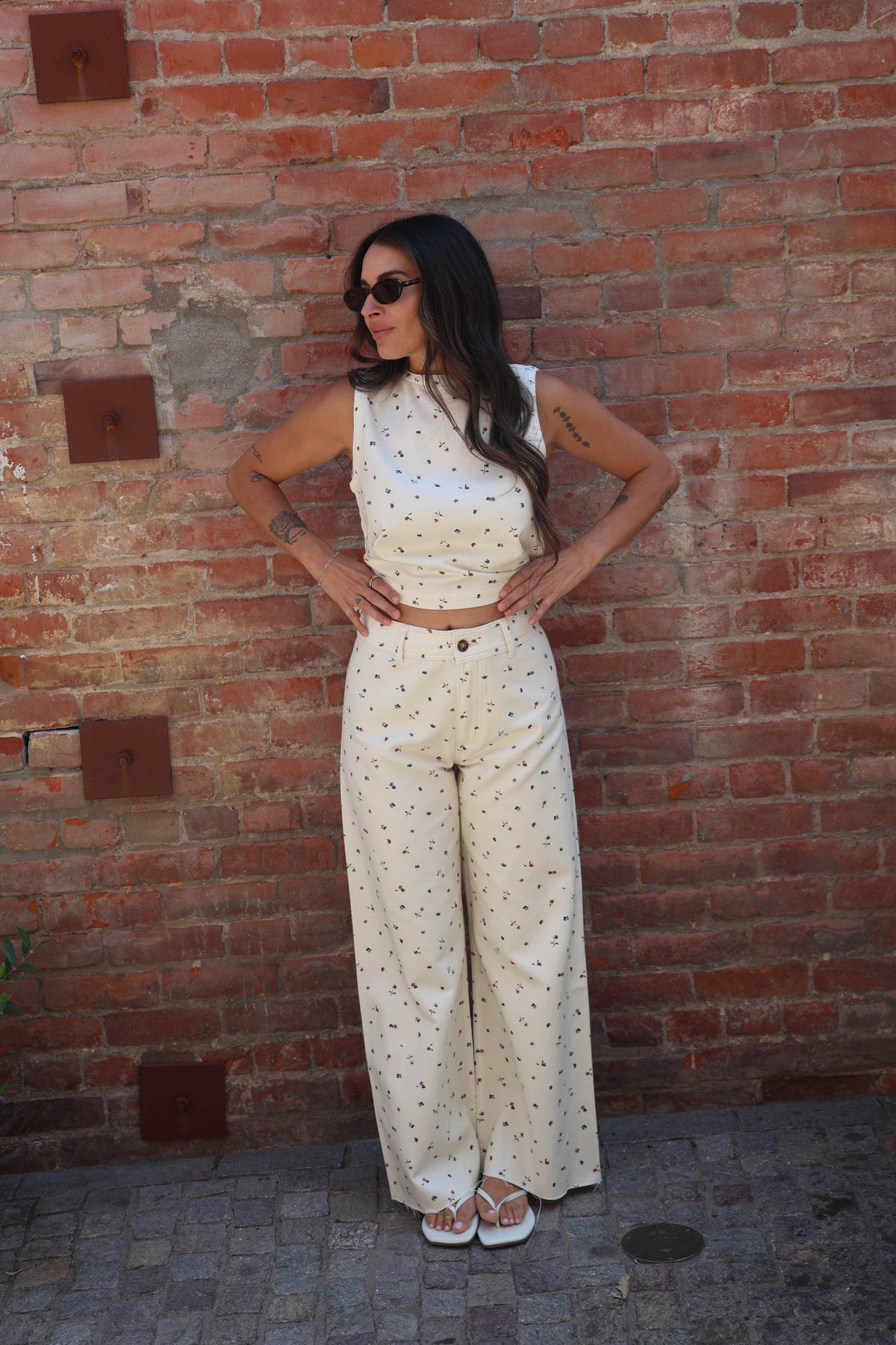 A woman wearing a matching sleeveless crop top and wide-leg pants set in cream with small black polka dots, standing against a red brick wall.