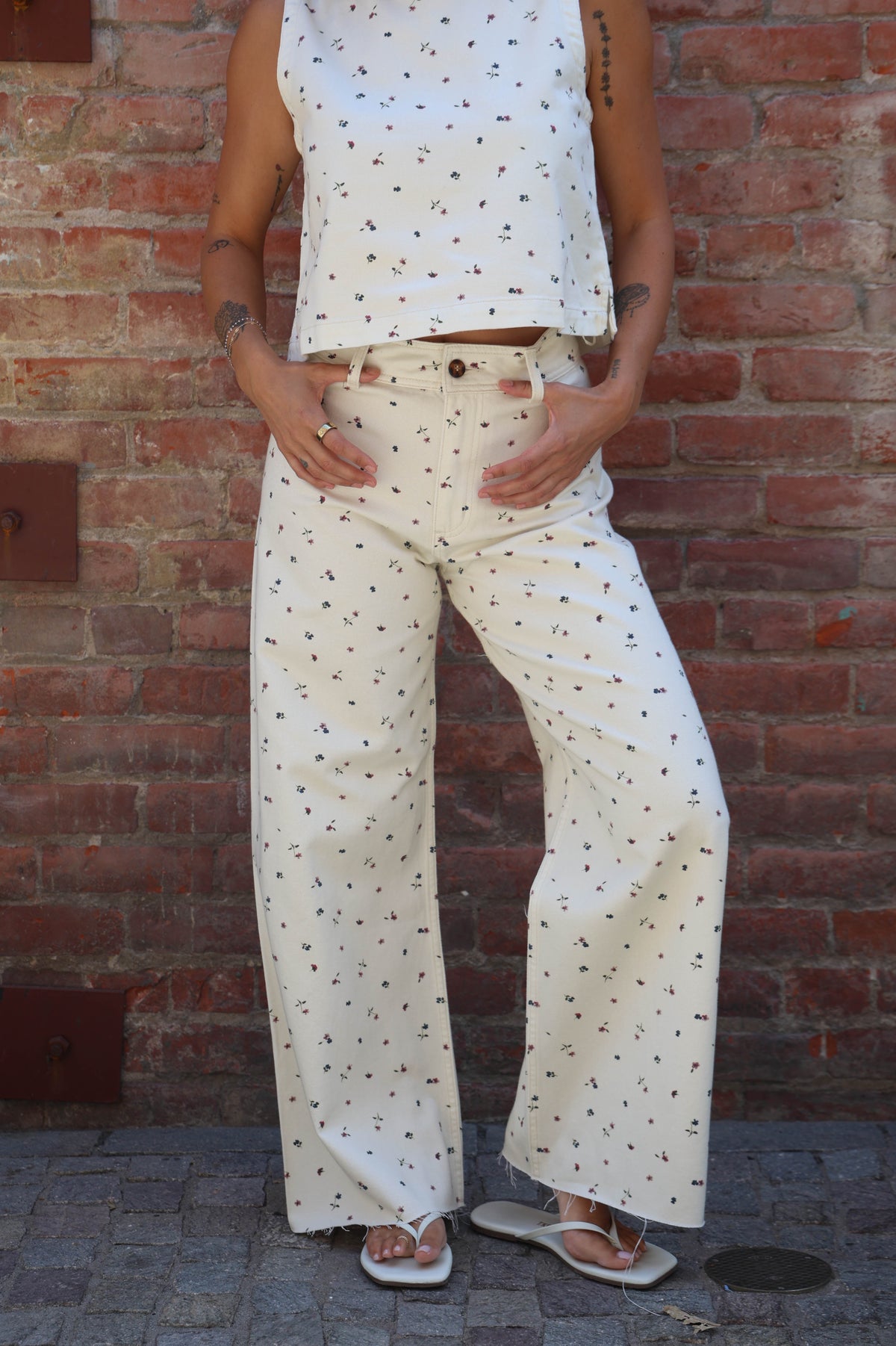 A person wearing a matching sleeveless crop top and wide-leg pants set in off-white with a small black polka dot pattern, standing against a red brick wall.