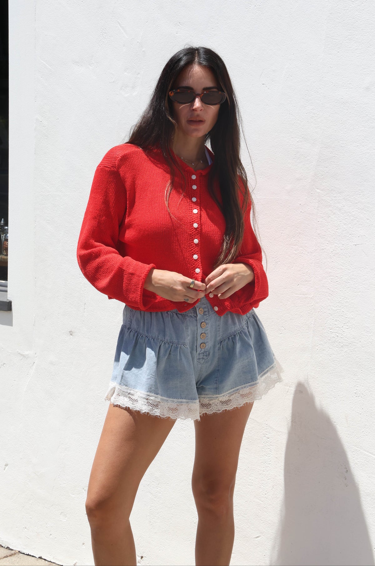 Woman wearing a red cardigan and denim shorts standing against a white wall.