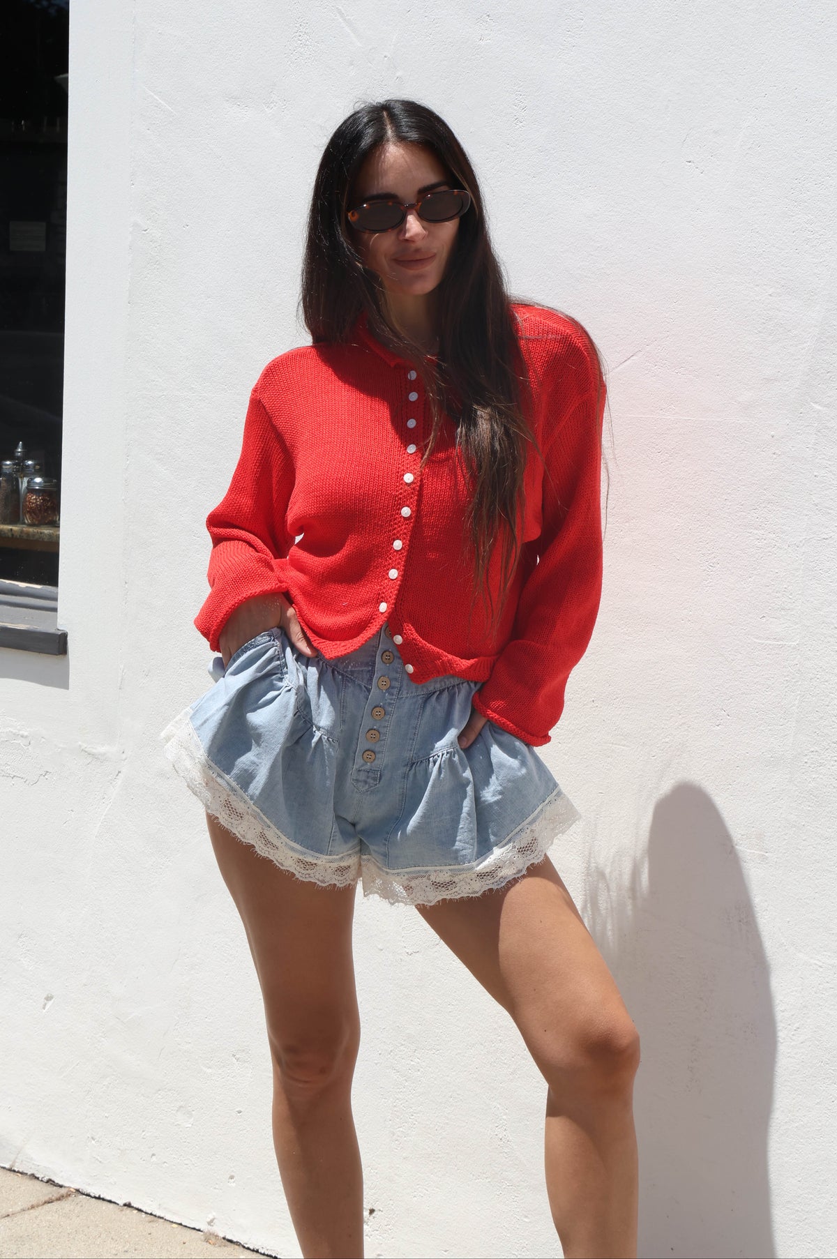 Woman wearing a red cardigan and denim shorts standing against a white wall.