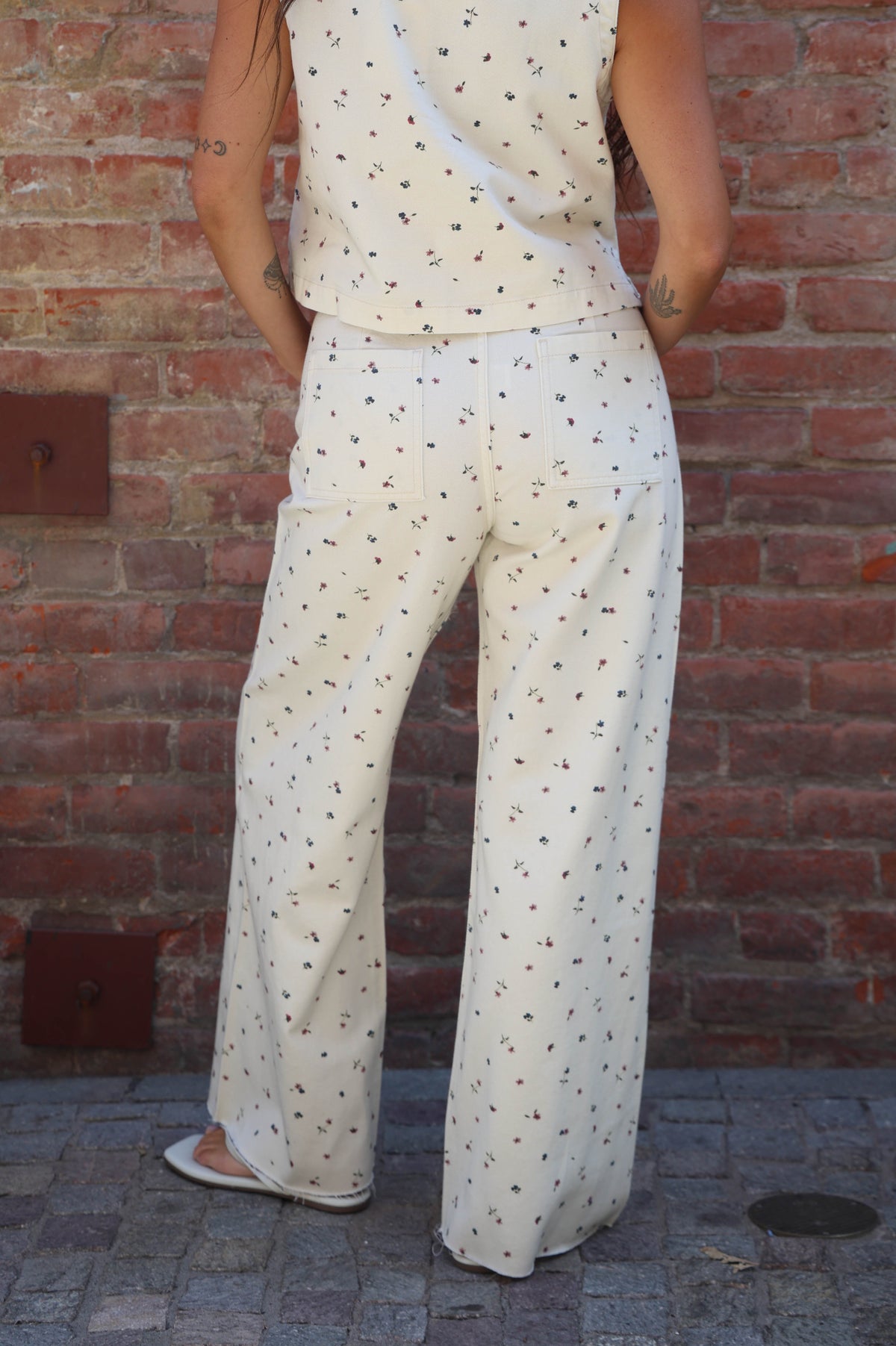 A woman shown from the back wearing a sleeveless wrap-style top and matching wide-leg pants in an off-white fabric with a small black geometric pattern, standing against a red brick wall.