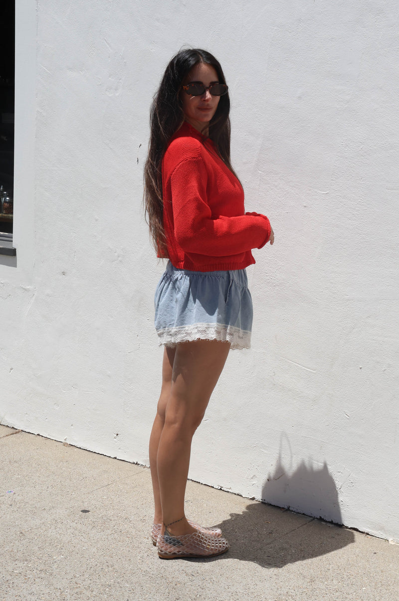 Woman wearing a red sweater and denim shorts standing against a white wall.
