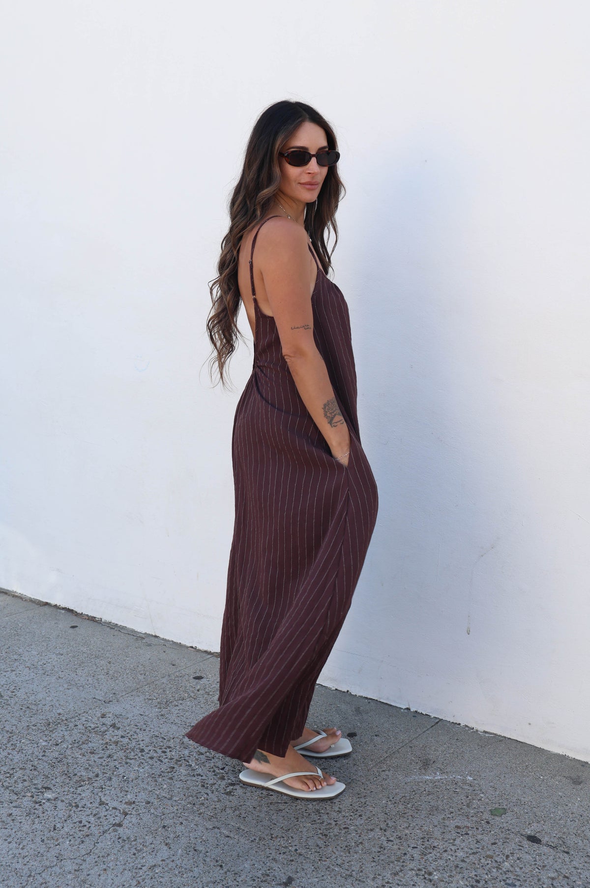 A woman wearing a dark brown ribbed maxi dress and sunglasses stands in profile against a white wall on a gray concrete ground.