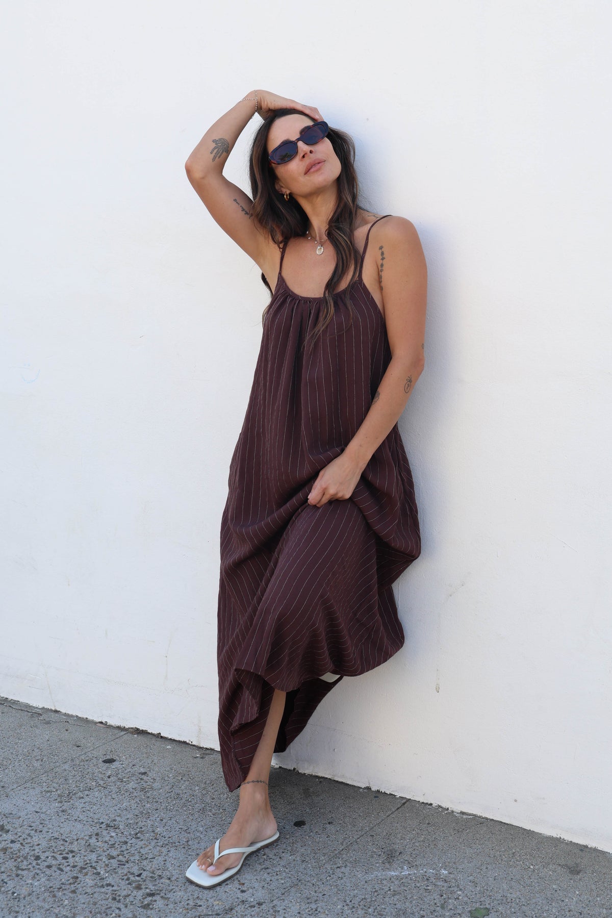 A woman wearing a dark brown pleated spaghetti-strap maxi dress and white flip-flops leans against a white wall on a concrete ground.