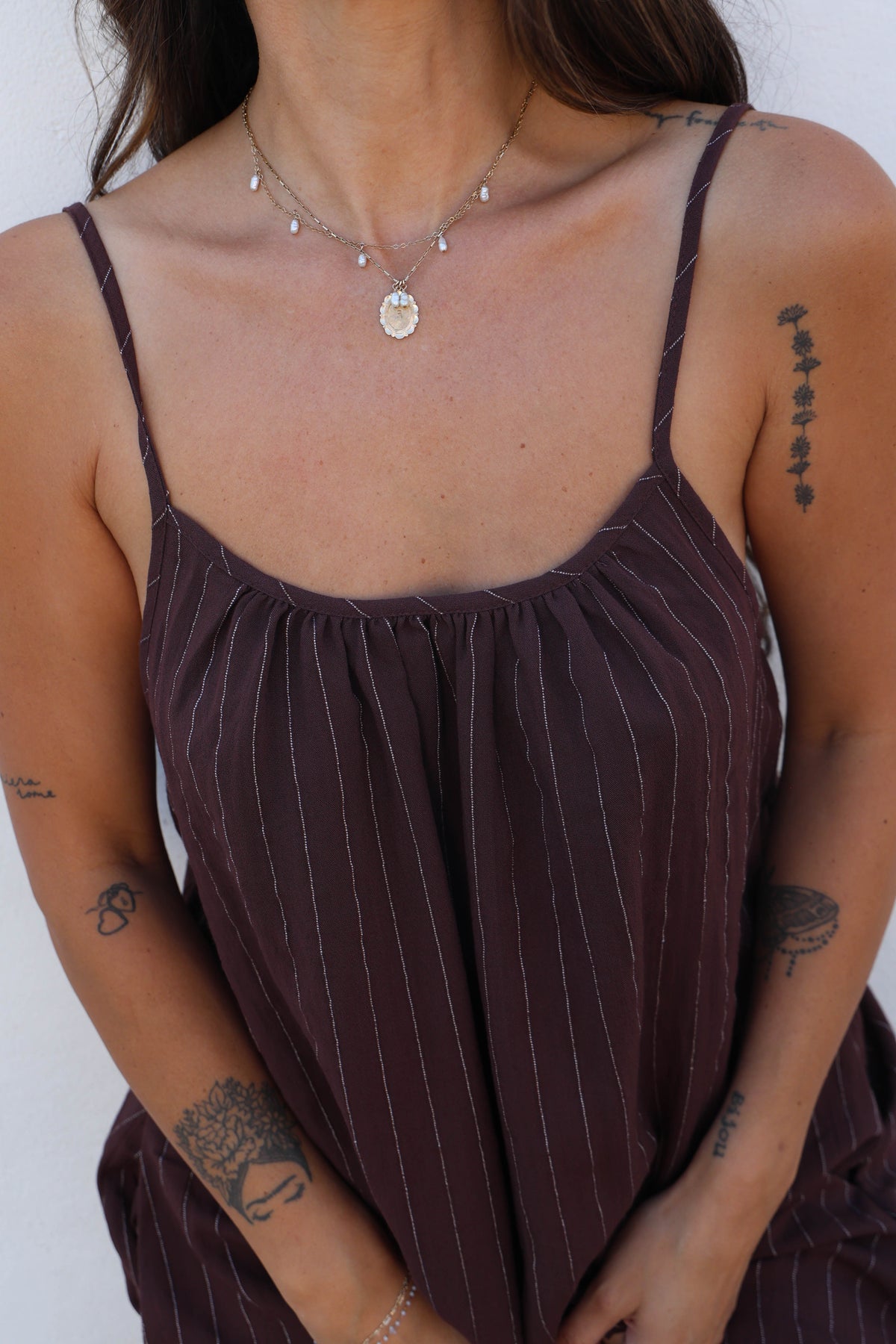 A close-up of a woman wearing a dark brownish-purple camisole top with thin vertical pinstripes and a gathered neckline, styled with a layered silver necklace.