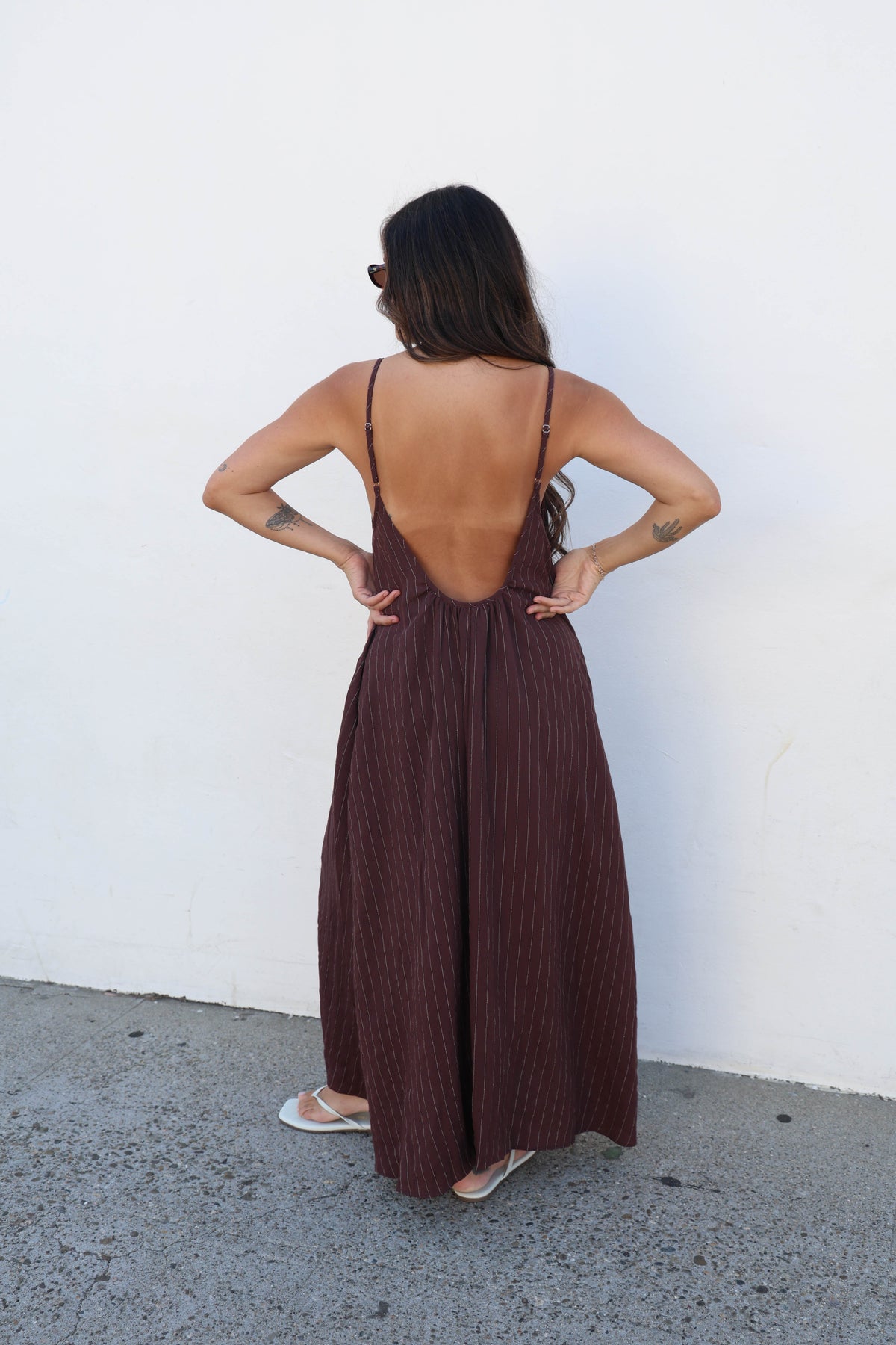 A woman viewed from behind wearing a dark brown open-back maxi dress with thin straps and a pleated texture, standing against a plain white wall.