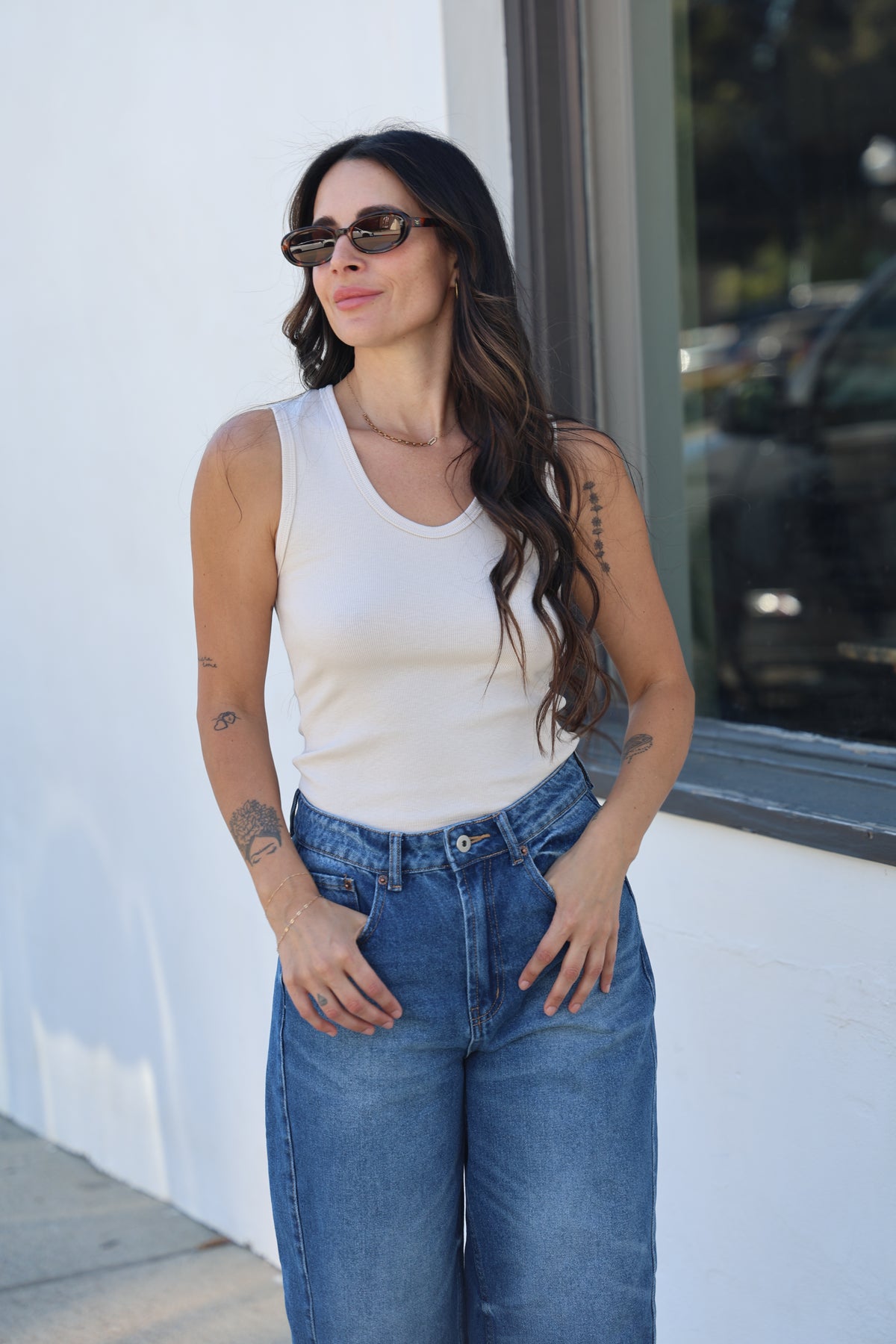 Woman wearing sunglasses and a white top with blue jeans, standing on a white surface.