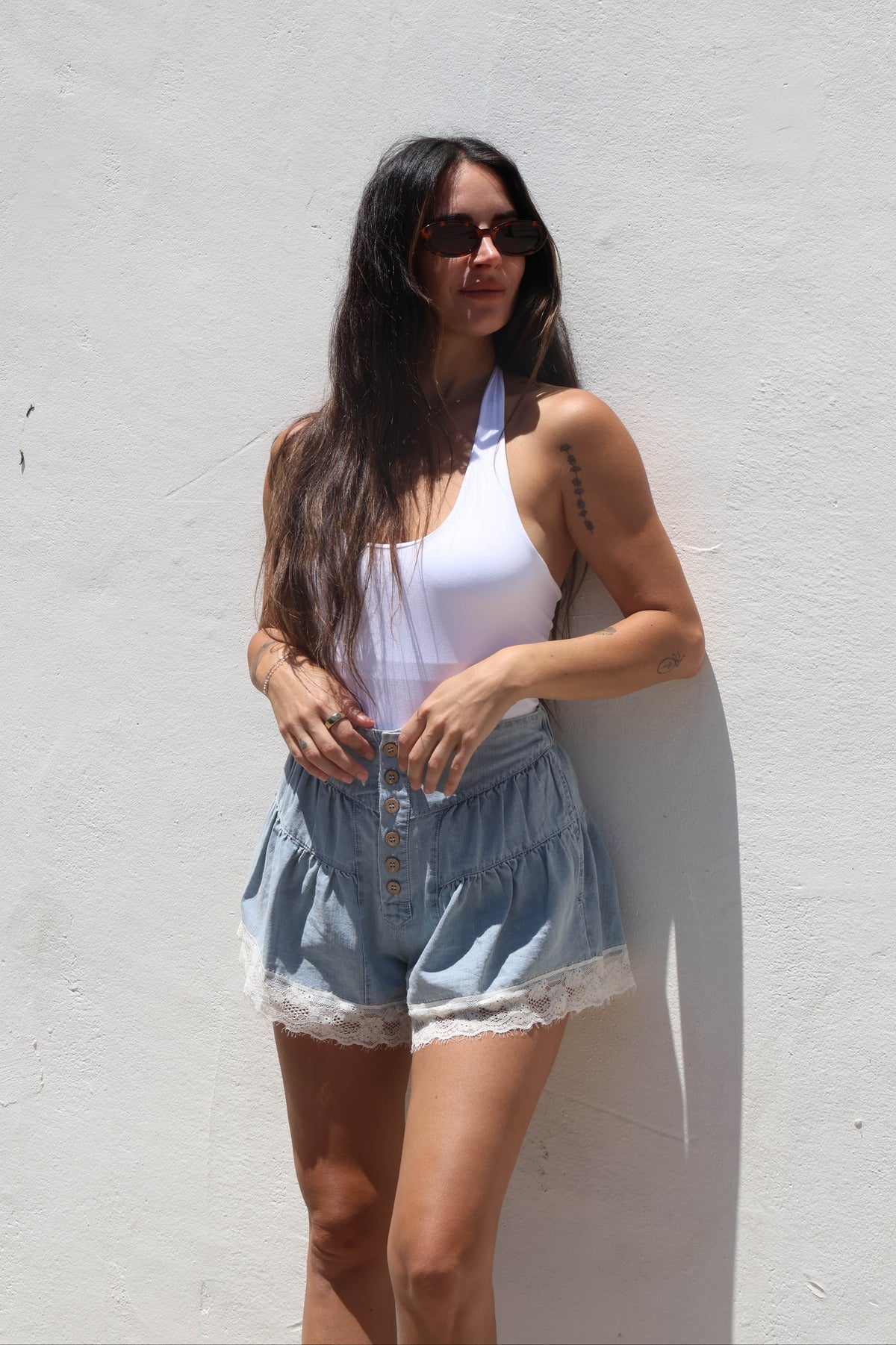 Woman wearing a white halter top and denim shorts against a white wall.