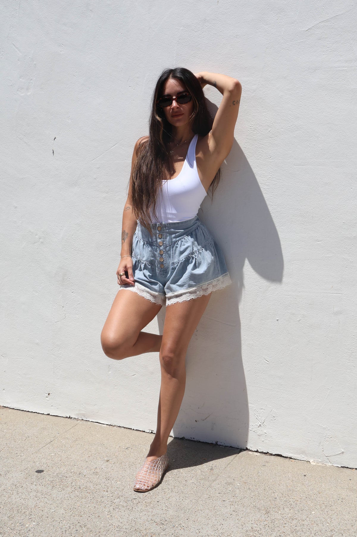 Woman in white tank top and denim shorts standing against a white wall.