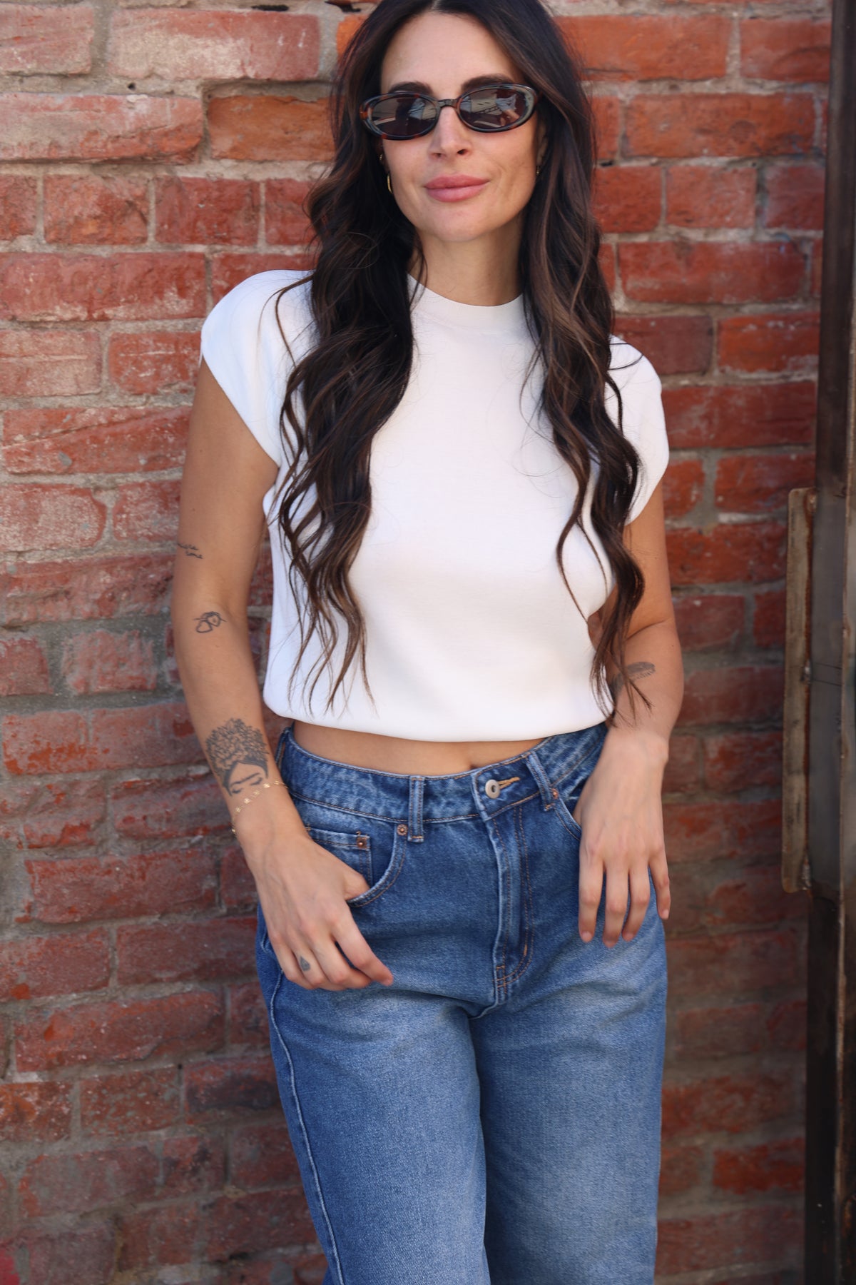 Woman wearing a white top and blue jeans against a brick wall.