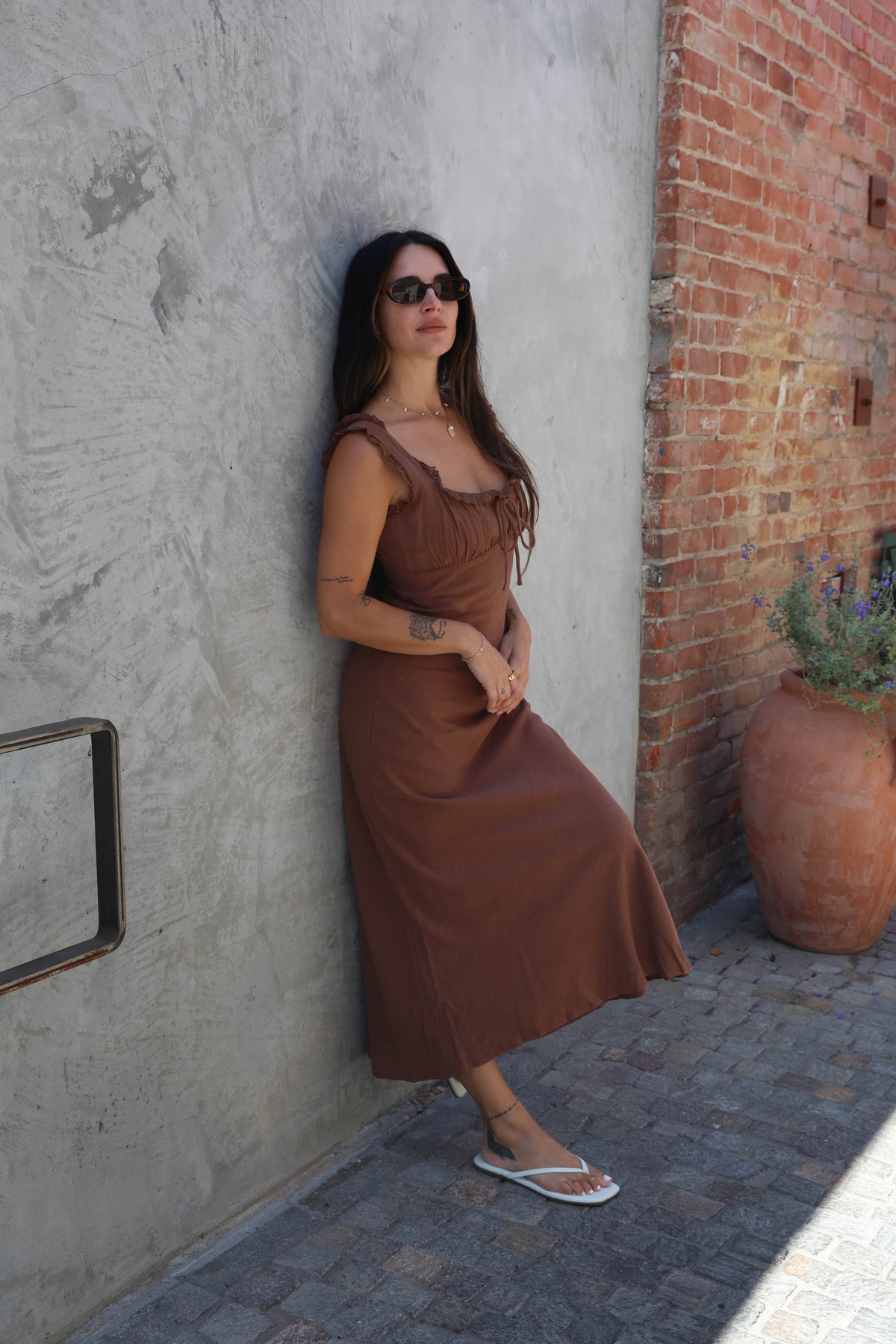 Woman in a brown dress leaning against a textured wall with a potted plant nearby.