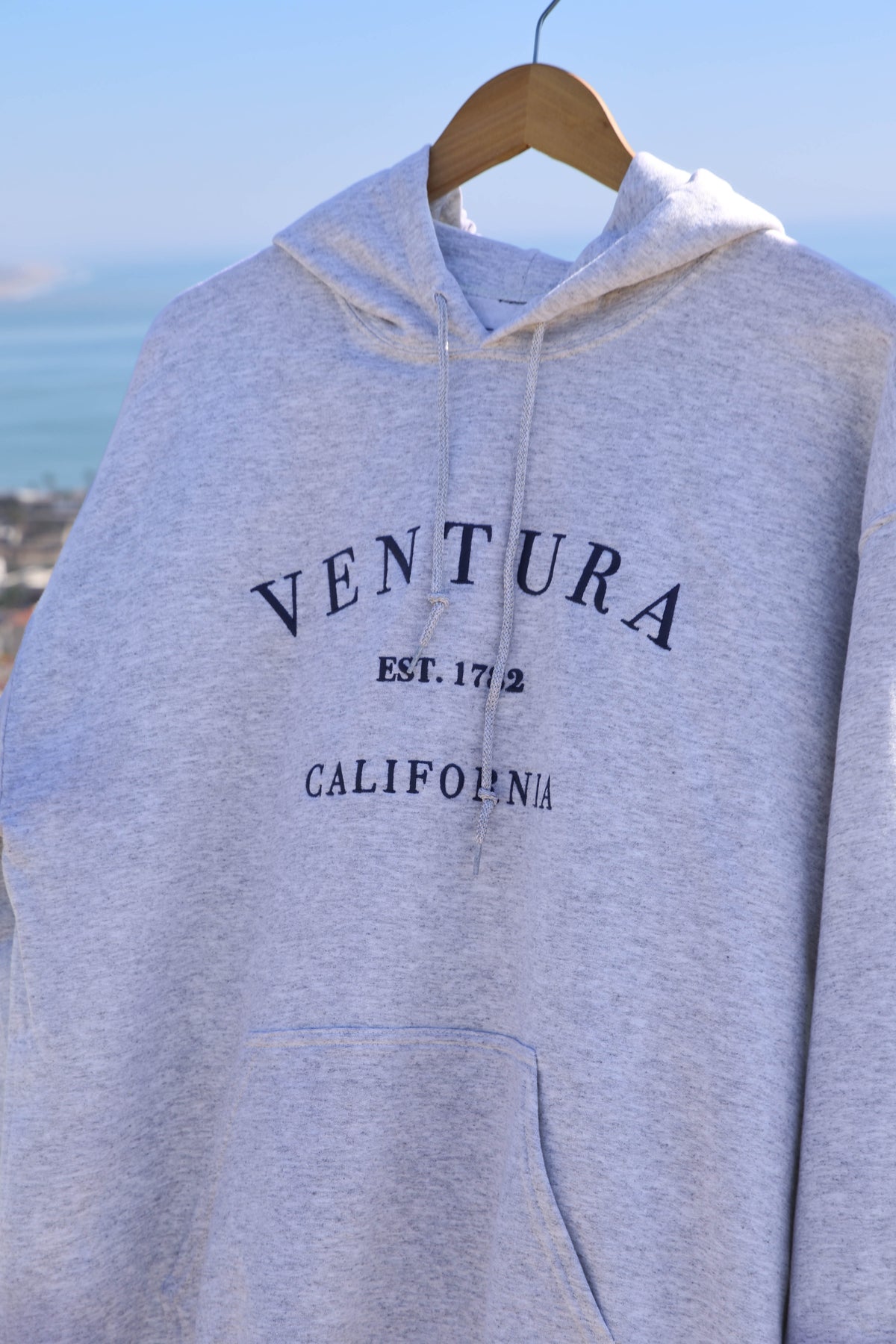 Gray hoodie with 'Ventura, California' text on a hanger against a scenic background