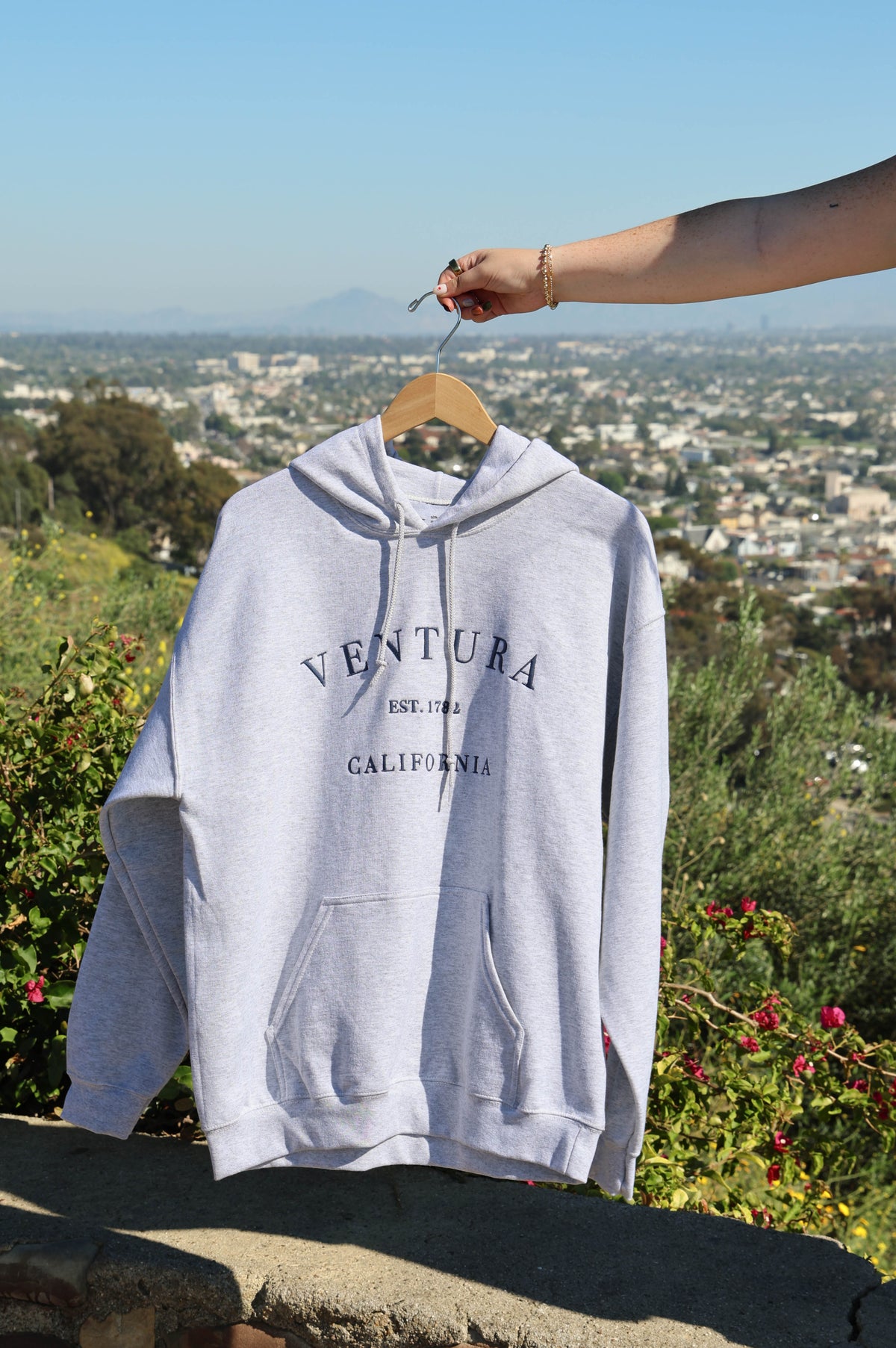 Gray hoodie with 'Ventura California' text on a hanger against a scenic backdrop