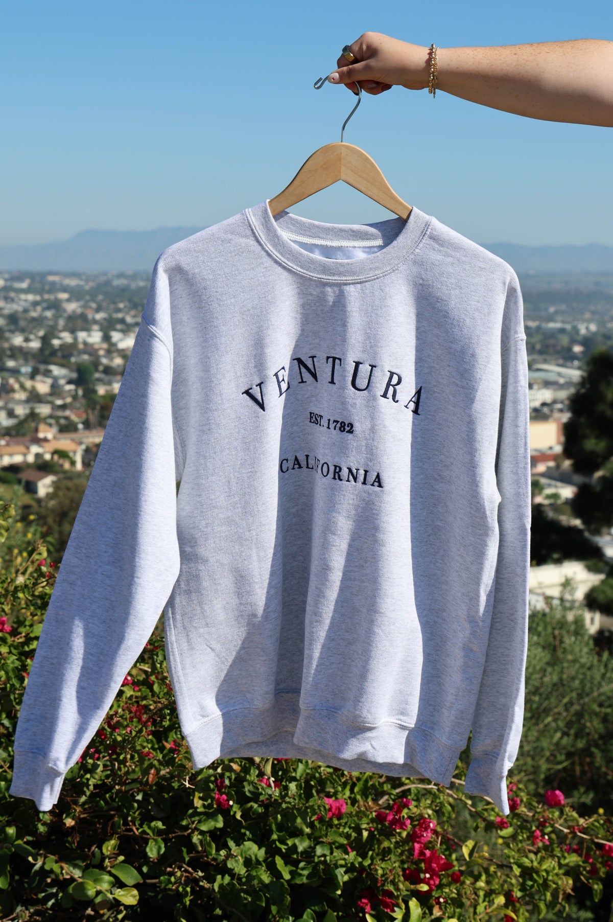 White sweatshirt with 'Ventura California' text on a hanger against a scenic background
