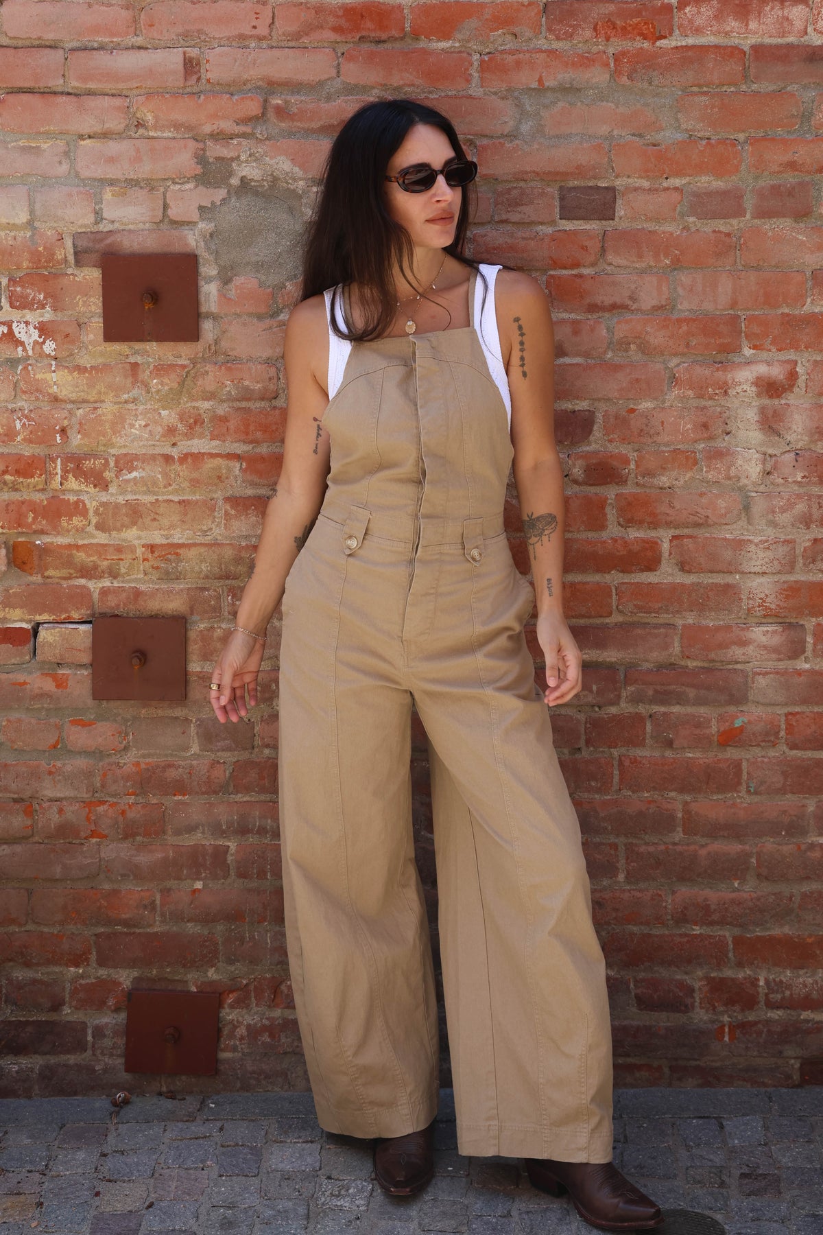 Woman wearing beige overalls standing against a brick wall.